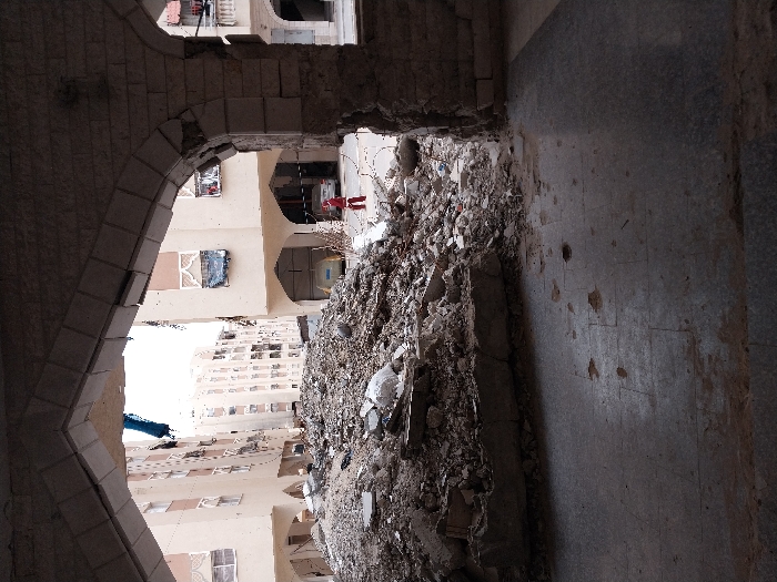 The image depicts a scene of extensive urban destruction in Khan Yunis, Palestinian Territory, viewed from within a damaged, light-colored stone archway. In the immediate foreground, a dark, tiled or paved floor is visible, marked with dirt, debris, and several small indentations or impact points. A large mound of concrete rubble, fragmented blocks, rebar, and construction debris dominates the central and left midground, rising significantly and obscuring the ground level beyond it.
Beyond this rubble, several multi-story, light-colored (beige/cream) buildings are visible in the background. These structures exhibit substantial damage, including shattered windows, exposed interiors, and hanging debris, such as a prominent blue fabric piece from an upper floor. The buildings feature arched entrances at ground level. To the right of the rubble pile and in the distance, a sole individual wearing a red top and dark trousers stands near a parked white sedan and a light-colored waste container. The sky above the buildings is bright and overcast. The environment shows widespread structural compromise and debris consistent with conflict damage.