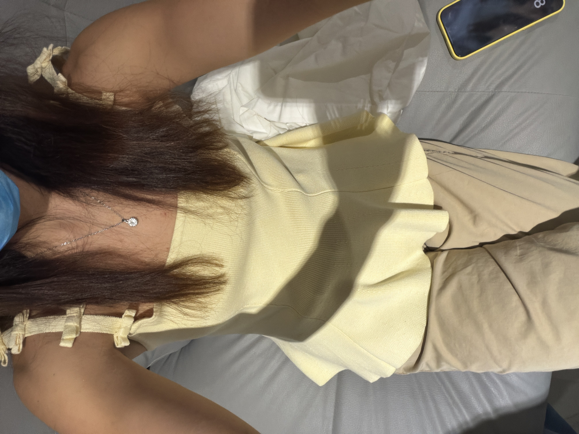 A woman is sitting indoors on a gray upholstered sofa. She is wearing a light yellow strapless top with tie details on the shoulders and tan trousers. A silver necklace with a small pendant is visible. A white cloth or fabric is beside her, along with a smartphone with a black screen and a yellow case. The location is Villahermosa, Mexico.