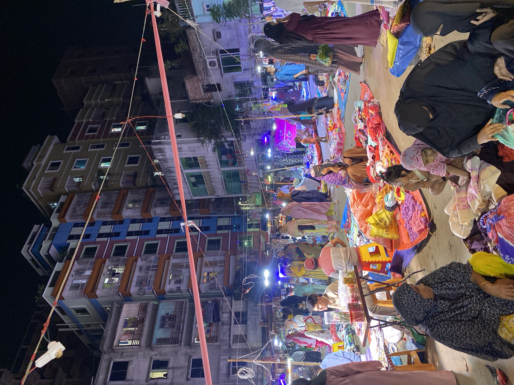 This media file captures a vibrant and crowded night market scene in Dhaka, Bangladesh. The setting is an urban street or alleyway, lined by tall, multi-story residential buildings that display various colors like blue, orange, red, and grey, with lights visible in many windows and balconies.
The time of day is clearly night, as indicated by the dark sky and the reliance on artificial lighting, which includes bare bulbs strung overhead and brighter blue lights illuminating market areas. The market itself is bustling with people, predominantly women and children, engaged in shopping and selling activities. Many individuals are dressed in traditional garments, including hijabs.
Goods are laid out on tarps, blankets, and low tables on the ground, suggesting an informal market setup. These items appear to be textiles, clothing, and possibly household goods. One vendor is seen with clear containers holding what seem to be red sweets or small items. A yellow bag with the text "FASHION TARGET" is visible among the merchandise.
Overhead, a complex web of electrical wires and temporary poles supports the market's lighting and other utilities. The atmosphere is lively and commercial, reflecting a common scene in densely populated urban areas. The perspective is somewhat tilted, emphasizing the height of the buildings and the spread of the market below.