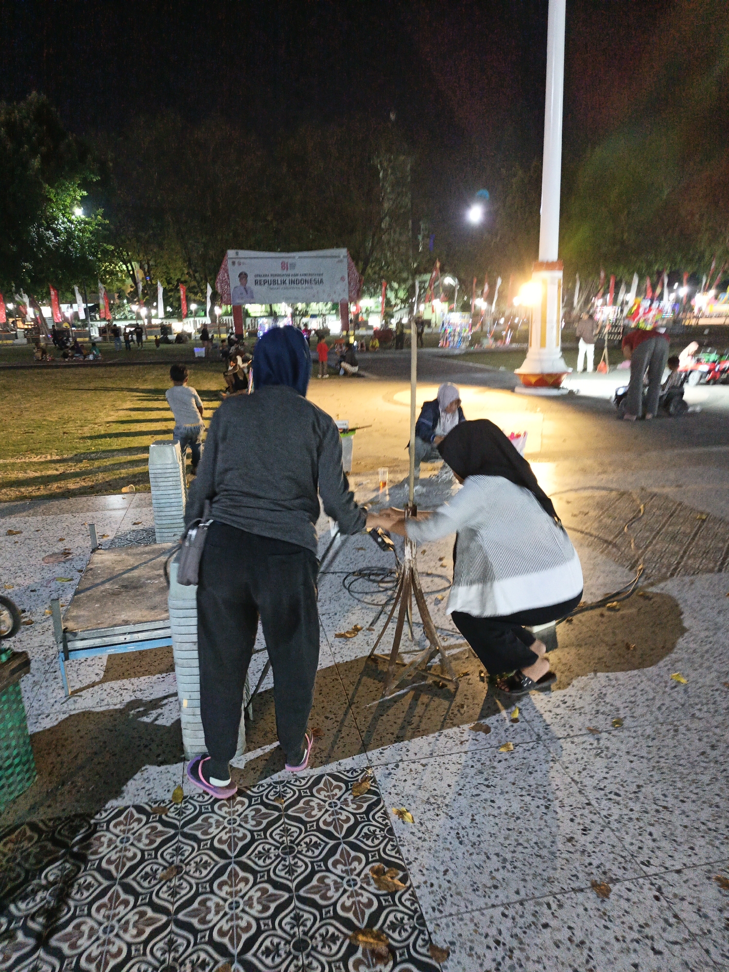 Two individuals are engaged in setting up equipment, likely for a performance or event, in an outdoor urban park at night. One person, facing away from the camera and wearing a dark hijab and a striped long-sleeved shirt over black pants, is crouched down, appearing to adjust a tripod. The other person, also facing away and wearing a dark blue hijab and a gray long-sleeved shirt over black pants, stands nearby, holding a cable or rope.
The scene appears to be in a public park, possibly a town square, judging by the presence of a large flagpole with a light fixture at its base, and a banner in the background with the text "REPUBLIK INDONESIA" and a portrait of a man. The ground is paved with patterned tiles and speckled concrete. Trees are visible in the background, illuminated by artificial lights. Other people are scattered in the background, some seated and some standing, suggesting a gathering or event is taking place or about to begin. The overall ambiance is that of a community event or festival in Klaten, Indonesia.