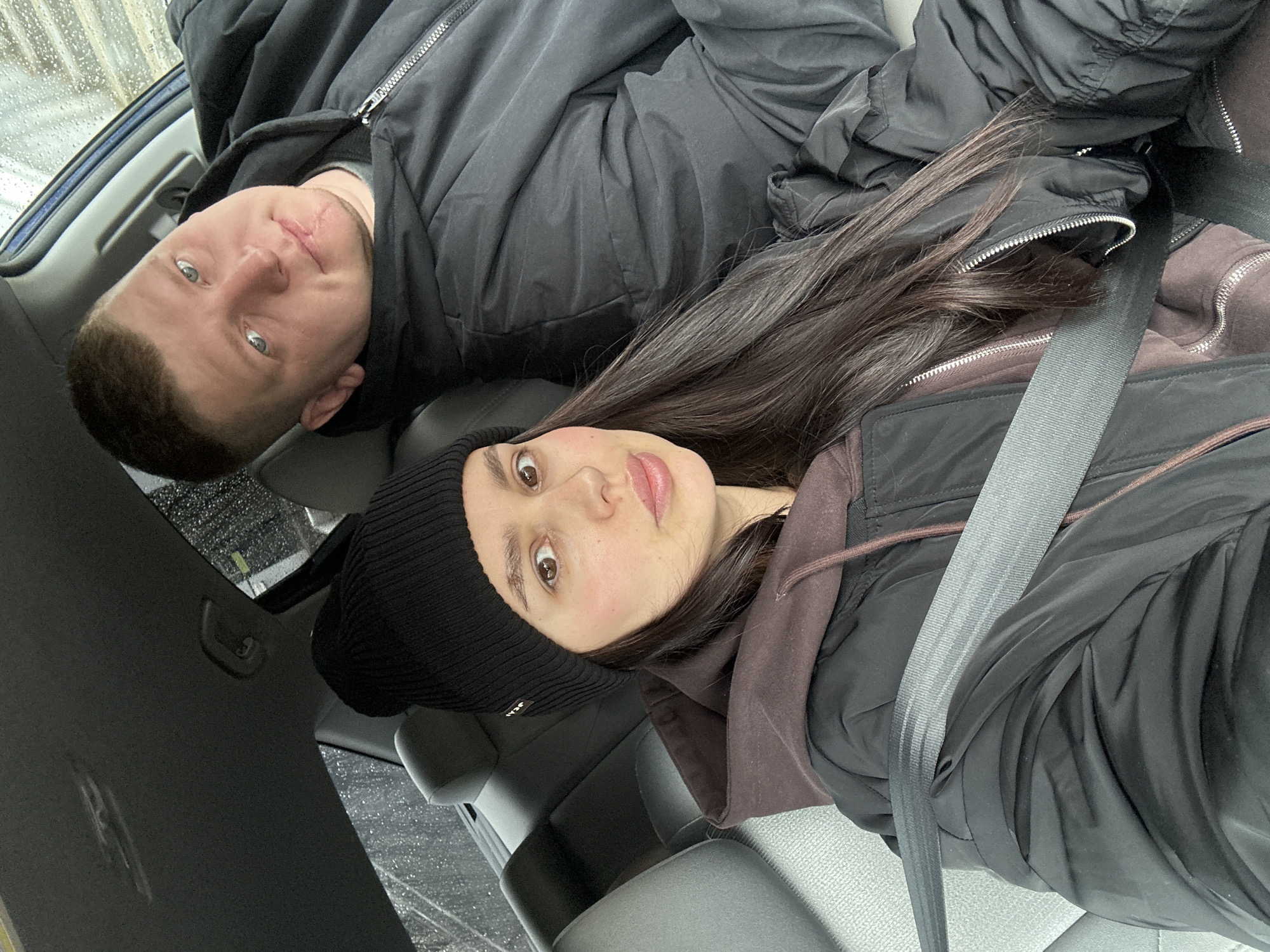 Two people are in a car. A man with short brown hair and blue eyes is wearing a black jacket. A woman with long dark hair and brown eyes, wearing a black beanie and a brown hoodie under a black vest, is also in the car. Both individuals are wearing seatbelts. Raindrops are visible on the car windows. The location may be in Chaikovsky, Russia.