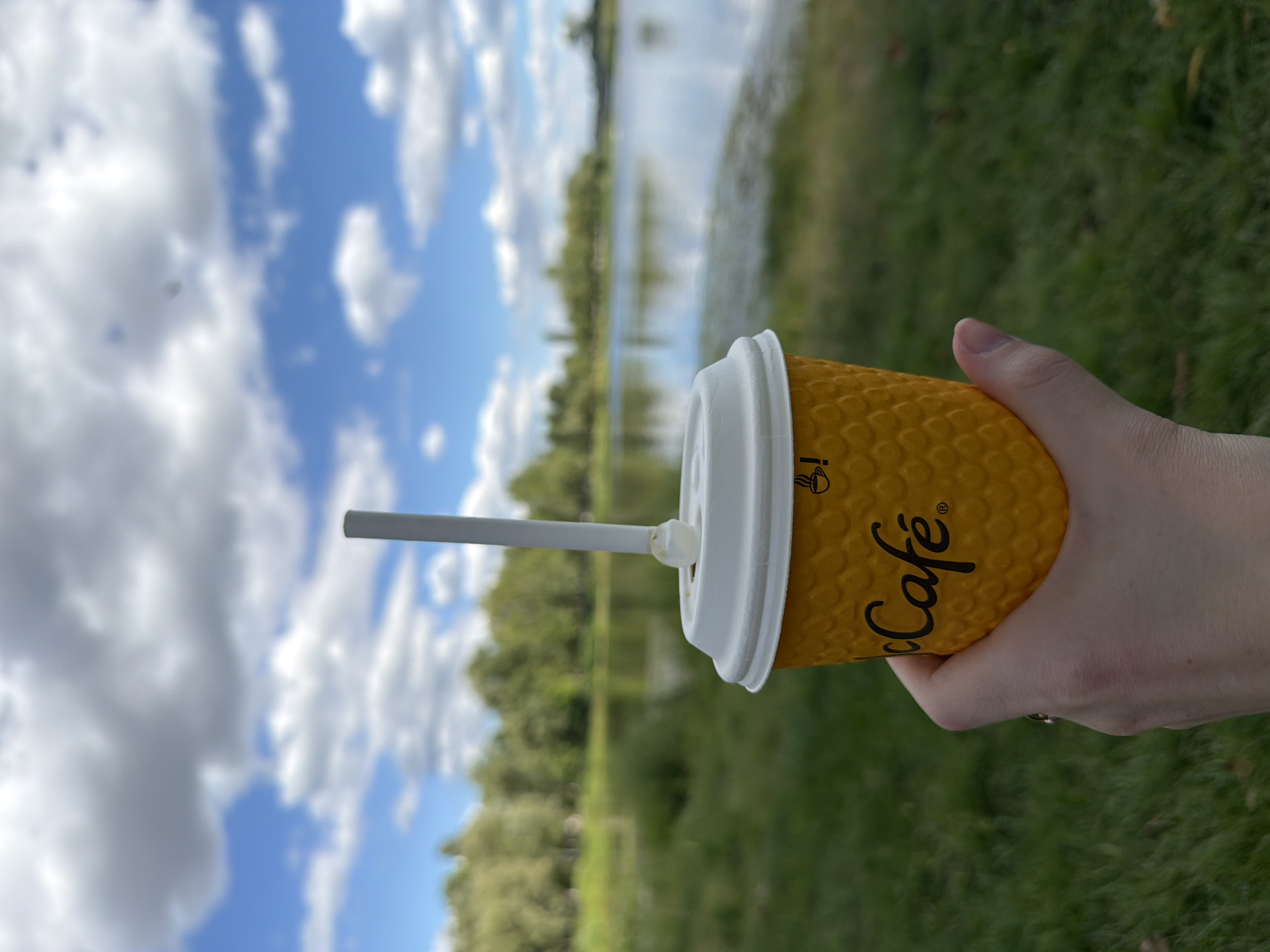 In Vyshhorod, Ukraine, a hand holds a yellow McCafe coffee cup with a white lid and a white straw. The cup has a textured honeycomb pattern and the McCafe logo in black. In the background, a body of water reflects a blue sky with scattered white clouds. Trees line the far bank of the water, and green grass covers the foreground. The lighting suggests it is daytime.