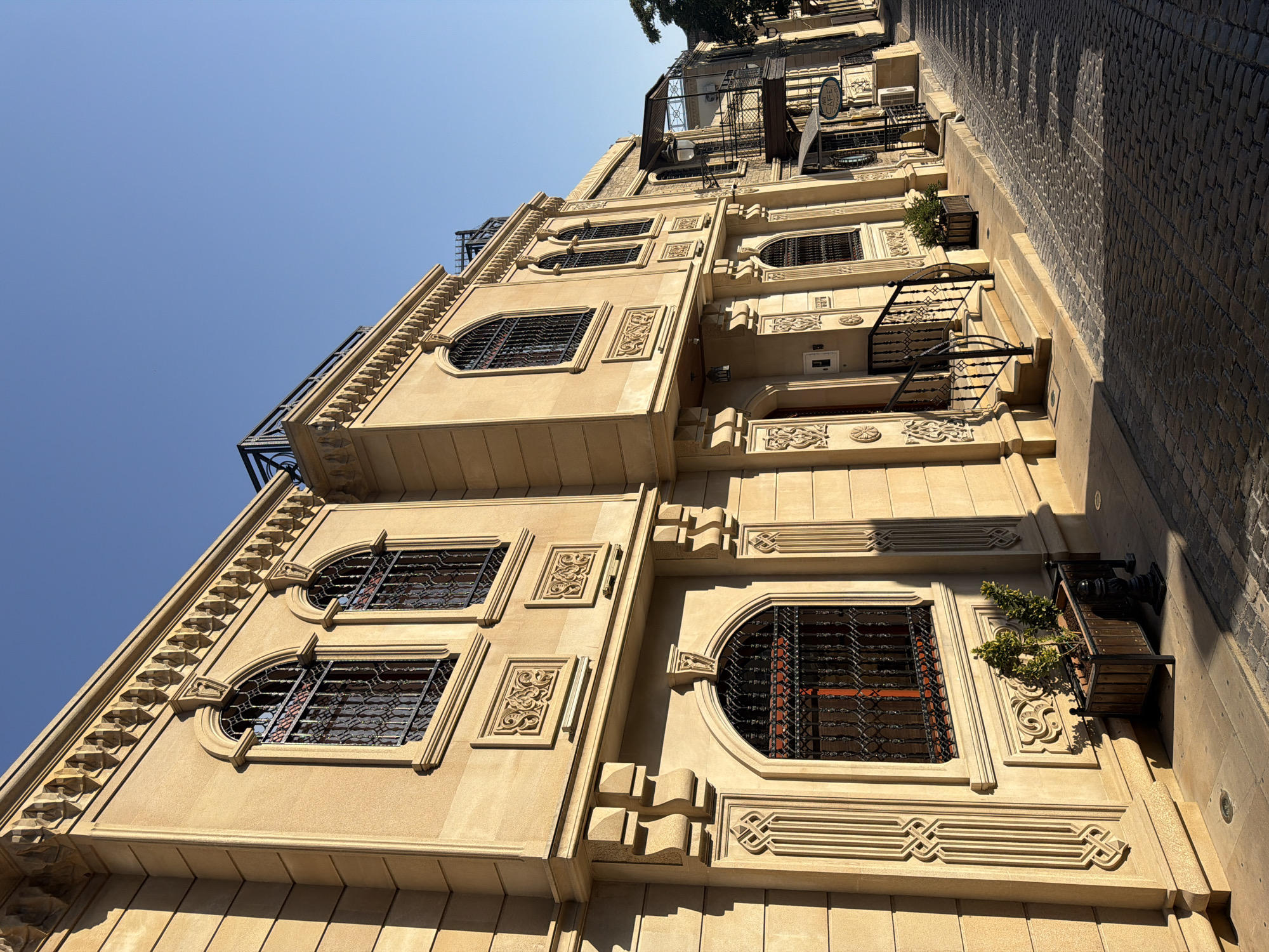 The image shows a sunlit cobblestone street in Sabail Raion, Azerbaijan, lined with a row of ornate, light-beige stone buildings. The architecture is characterized by arched windows with decorative iron grilles, intricate stone carvings above and below the windows, and a distinctive dentil molding along the roofline. The street itself is paved with uneven cobblestones, casting distinct shadows from the buildings and a nearby tree.
In the foreground, a planter box with a small, leafy green plant sits on the sidewalk next to a set of stairs leading to the entrance of one of the buildings. The entrance features a dark metal railing and a closed door. Further down the street, a cafe sign is visible on another building, along with an awning and a metal balcony. The sky is clear and blue, indicating a sunny day. There are no people visible in the image. The overall scene depicts a quiet, historic urban environment.