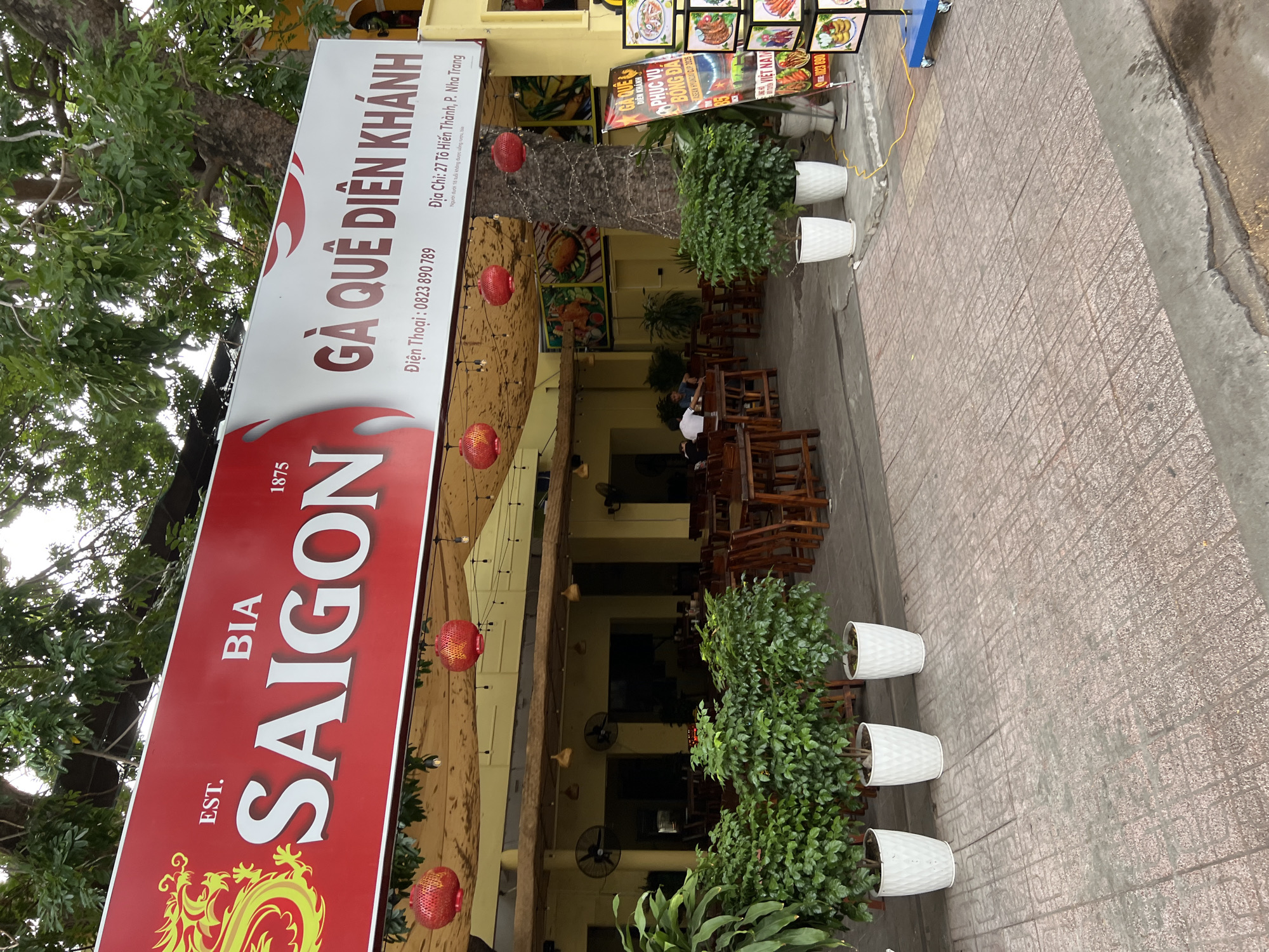A sign for "BIA SAIGON" and "GA QUE DIEN KHANH" is displayed above the entrance to a restaurant. The sign also lists the phone number 0823 890 789 and the address "27 To Hien Thanh, P. Nha Trang". Red lanterns hang from the ceiling over the outdoor seating area, where wooden tables and chairs are set up. Several large green potted plants are placed in front of the building. In the background, people are seated at tables inside the restaurant. A menu board with pictures of food is visible to the right of the entrance. The restaurant appears to be located on a city street with a tiled sidewalk.
