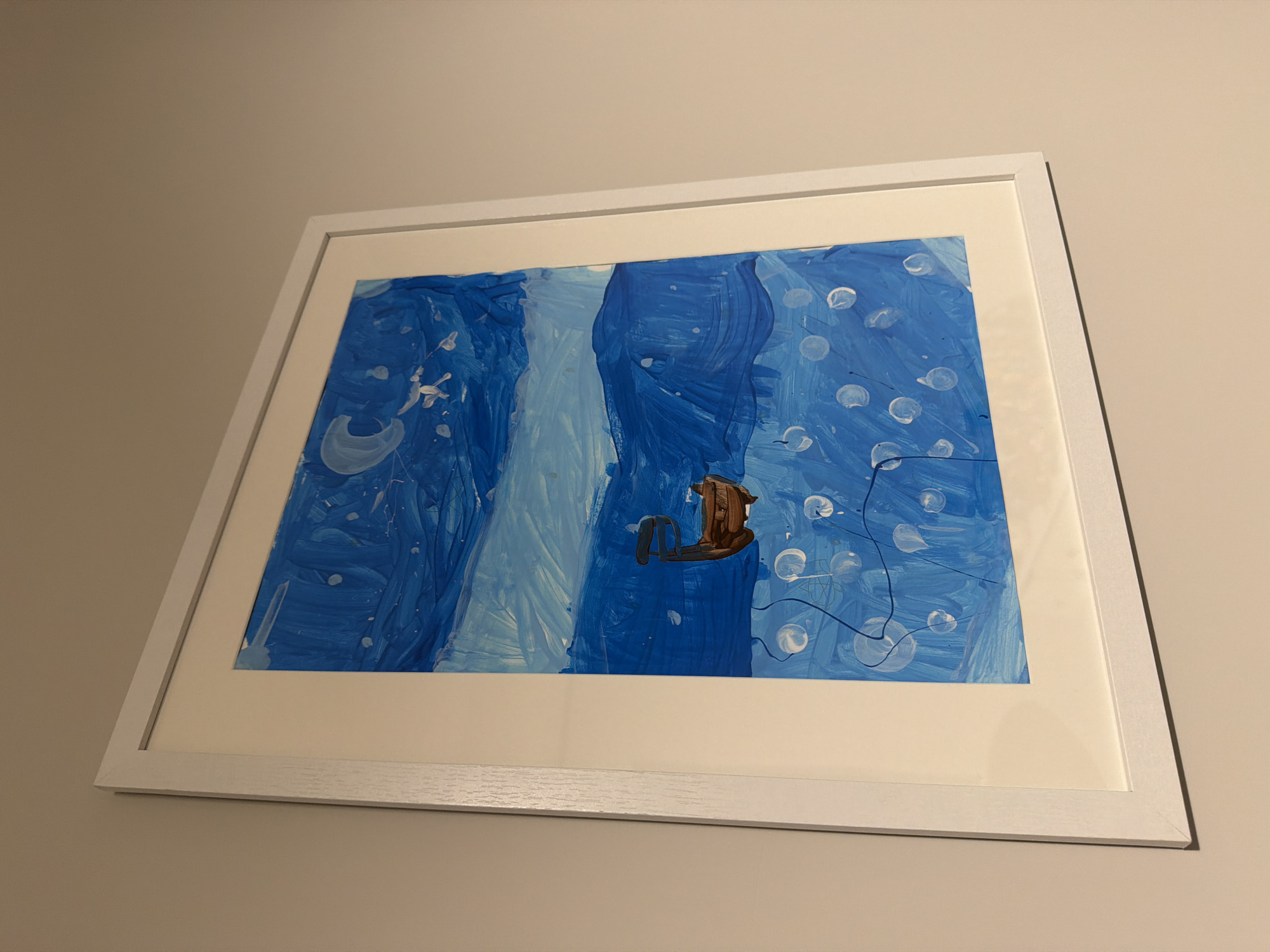 A framed painting hangs on a light beige wall. The frame is white and rectangular. The painting within the frame depicts a stylized waterfall in shades of blue, with a brown, object resembling a rocking horse positioned in front of it. The painting appears to be executed in thick brushstrokes of paint. White circular shapes, possibly representing bubbles, are scattered across the blue background. The perspective is angled slightly upwards toward the framed artwork.