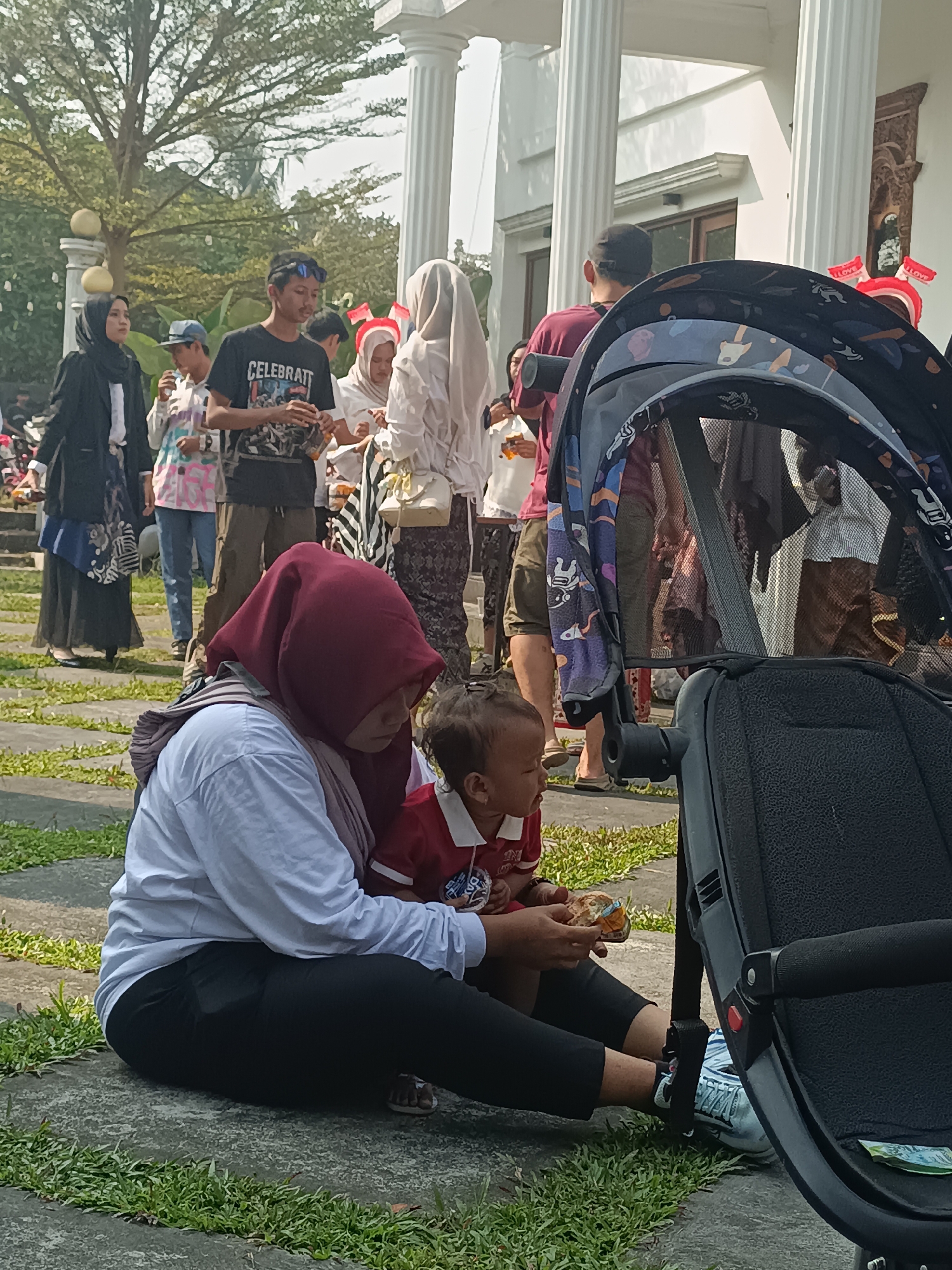 In an outdoor setting in Sleman, Indonesia, a group of people are gathered near a building with large white columns. In the foreground, a woman wearing a maroon hijab and a light blue long-sleeved shirt is sitting on the ground, leaning forward. She is wearing black pants and her feet are visible. Beside her, a young child in a red shirt is also seated, facing the woman. The woman appears to be offering a small food item to the child. Partially obscuring the background is a black stroller. In the mid-ground, several other individuals are visible, some wearing hijabs and traditional clothing, while others are in casual attire. One person is wearing a black t-shirt with the word "CELEBRATE" printed on it. The ground is paved with grey stone interspersed with patches of green grass. The environment suggests a daytime gathering or event.