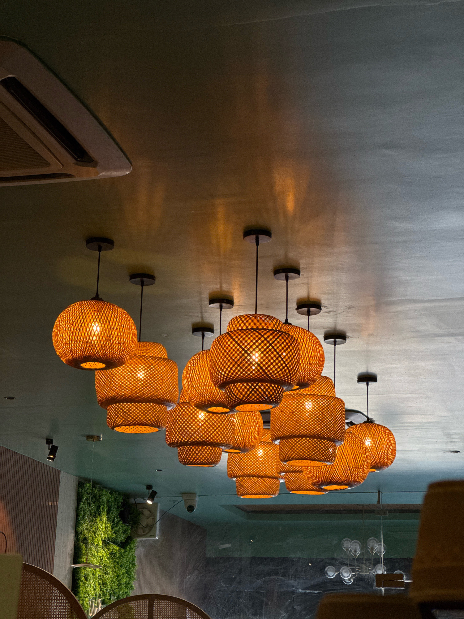 The image captures a close-up view of a cluster of woven, orange bamboo pendant lamps hanging from a textured, dark green ceiling. The lamps emit a warm, soft glow that casts intricate shadows on the ceiling. To the left, a portion of a white air conditioning unit is visible. Further to the left and down, a section of a wall with vertical wooden slats can be seen, adjacent to a living green wall. In the background, a security camera is mounted on the ceiling. The scene appears to be an indoor setting, possibly a restaurant or cafe, with furniture elements, including a part of a woven chair, visible in the foreground and lower right corner. The overall ambiance is warm and inviting, with a focus on natural materials and ambient lighting. The image does not contain any people, and no specific details suggest a location in Hisar, India.