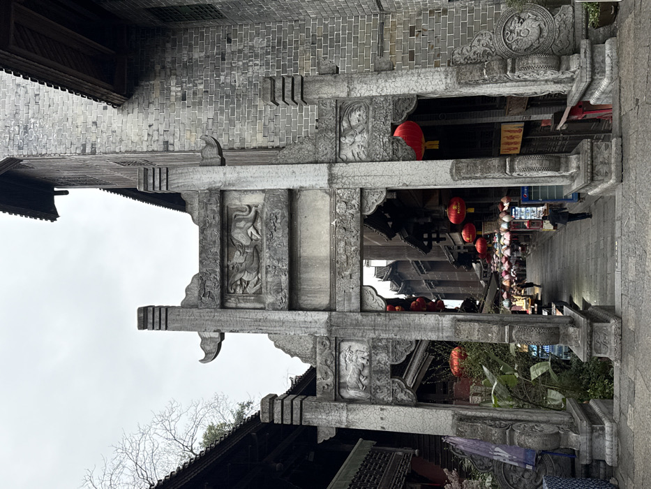 The image presents a stone archway (paifang) acting as an entrance to a historic street within Pengshui Miao and Tujia Autonomous County, China, under an overcast sky. The archway, constructed from weathered gray stone, features a central lintel with relief carvings of two stylized mythical creatures, possibly dragons, facing each other. Below this, smaller panels on either side of the archway display carved animal figures, potentially lions, amidst decorative patterns.
Beyond the archway, a stone-paved street, initially descending with steps, extends into the distance. It is flanked by multi-story buildings housing shops or stalls with traditional architectural elements. Numerous red spherical lanterns are suspended across the street overhead, continuing further down. A dense arrangement of colorful, tiered parasols or umbrellas also provides overhead cover further into the street. Several pedestrians are visible walking along the street, including one individual in a dark jacket walking away from the camera on the right side of the thoroughfare. A blue Pepsi branded refrigerator is visible on the right side of the street, just past the archway.
To the immediate right of the archway, a multi-story building constructed of gray brick with dark wooden eaves rises prominently. To the left, a darker building with traditional Chinese rooflines and visible dark wooden structural elements is partially observable. Bare tree branches are silhouetted against the gray sky in the upper-left quadrant, and green foliage grows near the base of the archway on the left side.