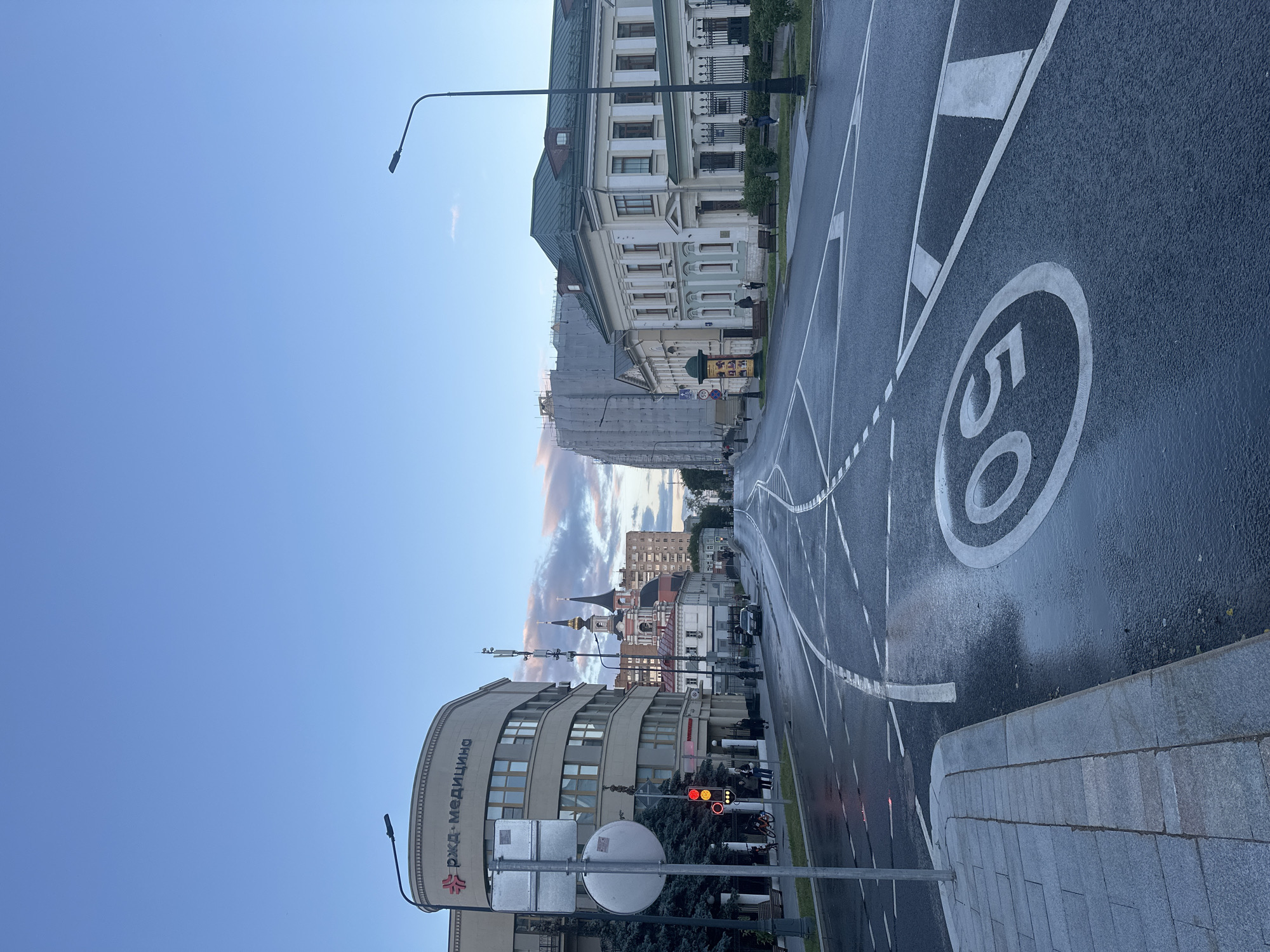 The image depicts a street scene in Moscow, Russia, likely in the late afternoon or early evening, given the soft lighting and overcast sky with hints of sunset colors. The wet asphalt suggests recent rain. The street is wide with multiple lanes and marked with various road markings, including a speed limit sign of "50" within a circle.
On the left side of the street, a modern, curved building with the Cyrillic inscription "РЖД-МЕДИЦИНА" (RZD-MEDITSINA) stands out. This is a medical facility associated with Russian Railways. Next to it is a traffic light, currently showing red and yellow. A pedestrian walkway with gray paving stones is visible along the sidewalk. Several people are walking on the sidewalk, and bicycles are parked nearby. Trees line the area in front of the building.
Further down the street, traditional Moscow architecture is visible. A church with a pink and white facade and two distinct domes is prominent. Adjacent to it are older, light-colored buildings with green roofs, one of which has ornate architectural details. In the background, taller, more modern buildings can be seen, one of which is covered in scaffolding, indicating ongoing construction or renovation. Streetlights are positioned along the road. A few cars are present on the street, and distant figures can be observed walking. The overall impression is of a bustling urban environment with a mix of modern and historic structures.