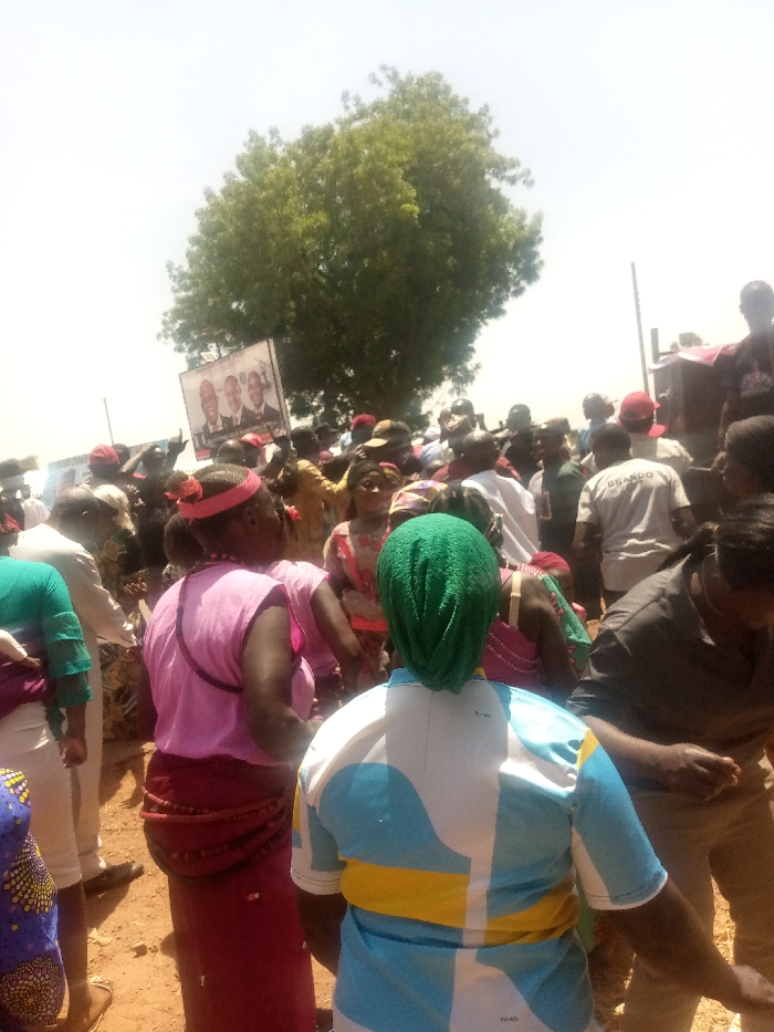 The image captures a densely packed outdoor gathering of numerous individuals in Kagoma, Nigeria, under a bright, sunny sky. The ground appears to be dry, reddish-brown earth. In the immediate foreground, facing away from the viewer, is a person wearing a green head covering and a short-sleeved shirt patterned with light blue, white, and yellow geometric shapes. To their left, another individual, also facing away, wears a pink sleeveless top and a red skirt or wrapped garment, adorned with a multi-strand beaded belt or waistline.
The crowd comprises primarily adults, many wearing diverse clothing including casual shirts, traditional wraps, and red caps. Prominently displayed and held aloft within the crowd is a white rectangular banner featuring three portraits of men. From left to right, the portraits show a dark-skinned man, a lighter-skinned man, and another dark-skinned man. Partial text is visible below the portraits. Further back, an individual is seen wearing a white t-shirt with the word "BRANDD" or "BRANNDO" printed on the back. In the upper center background, a large, full green tree stands against the bright sky. To the far right, indistinct structures and a vertical pole are visible. The people are congregated closely together, suggesting a public event or assembly.