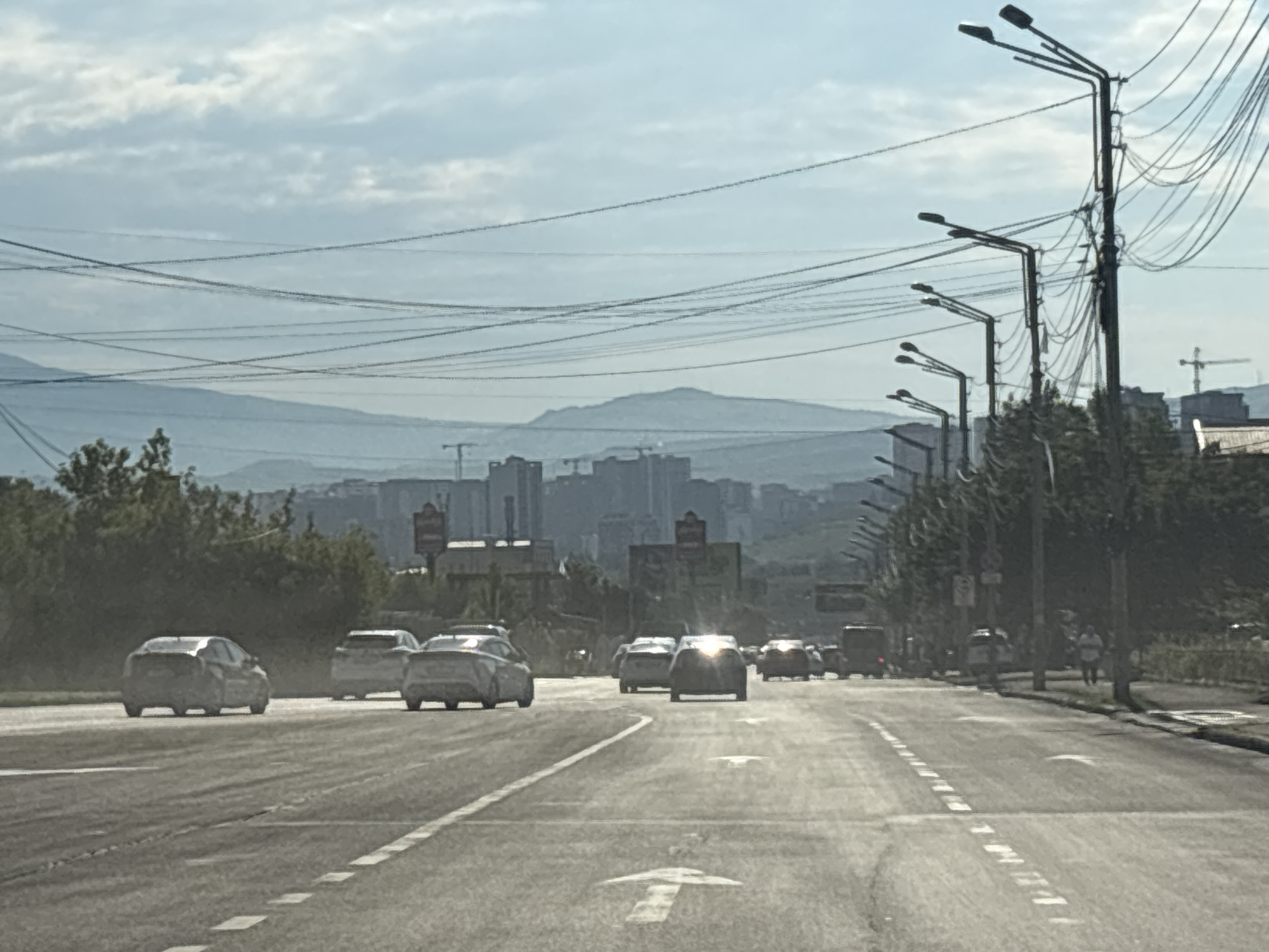 The image depicts a street scene in Tbilisi, Georgia, under a cloudy sky. Several cars are driving on a multi-lane road, with one car in the foreground appearing to be a white SUV. On the right side of the road, there are several tall streetlights and a complex network of overhead power lines. In the background, a distant city skyline is visible, characterized by several buildings and construction cranes, suggesting ongoing urban development. Beyond the city, a range of hills or mountains can be seen, softened by atmospheric haze. A few pedestrians are present on the right sidewalk, and trees line the side of the road. The overall impression is of a typical urban environment on a slightly overcast day.
