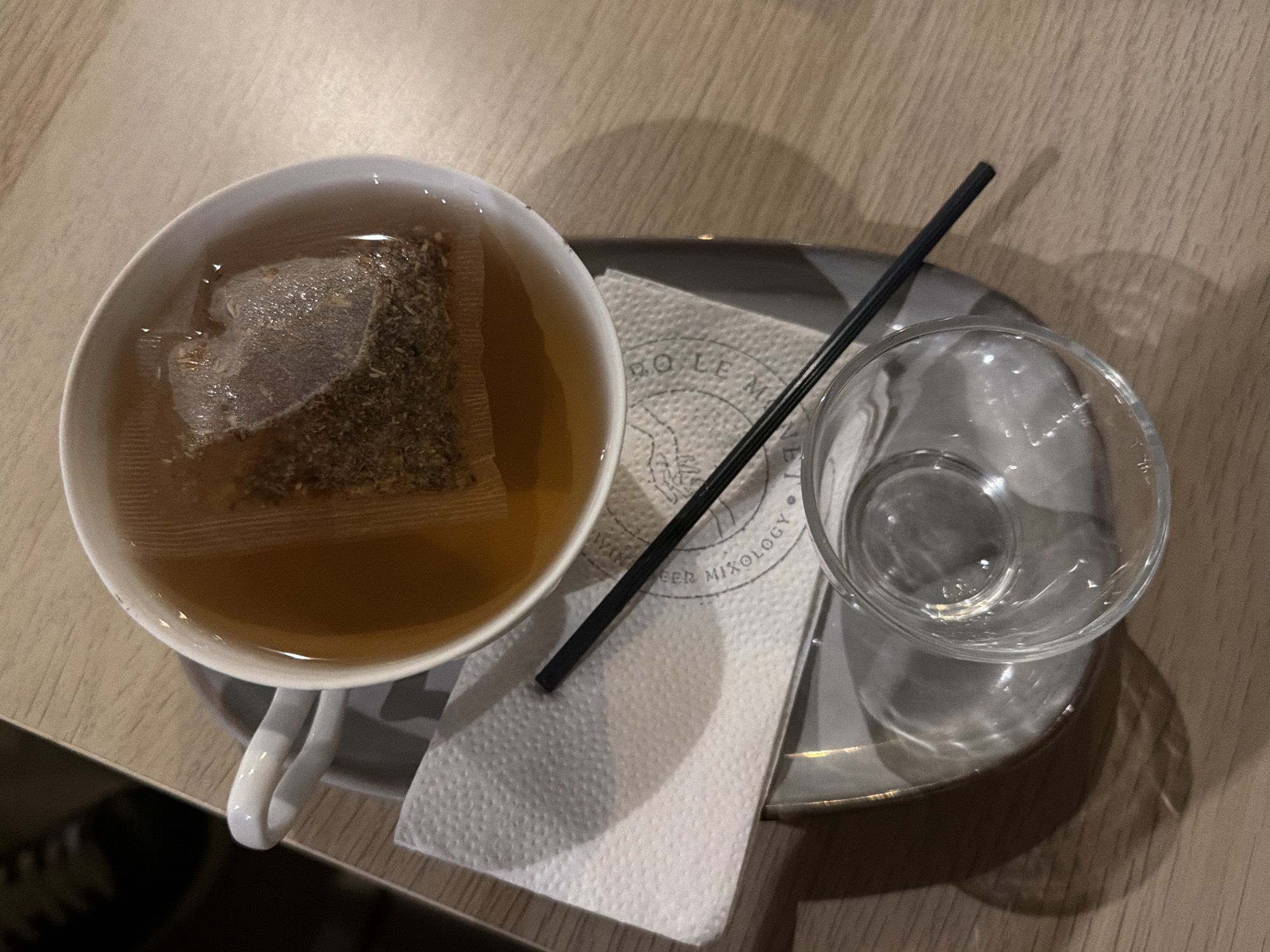 An overhead shot presents a hot beverage setup on a light wooden table, likely within a cafe or restaurant. A white ceramic teacup, filled with light amber-colored tea, occupies the left side of the frame. A pyramid-shaped tea bag, containing dark, finely ground herbs or tea leaves, is submerged in the hot liquid, actively steeping.
Beside the teacup, a small, dark grey or black tray holds a folded white paper napkin and a clear, empty glass. A slender black straw rests diagonally across the napkin. The glass, reflecting some light, is positioned to the right of the napkin.
Visible text on the napkin clearly reads "BO LEM" with "WINE BEER MIXOLOGY" underneath, indicating the name and services of the establishment. This suggests a casual, possibly evening, setting. Shadows from the cup, glass, and straw are cast across the table and tray, consistent with indoor lighting. There are no people visible, implying a quiet moment or an unoccupied table. The image captures the simple act of preparing or enjoying a cup of tea.
