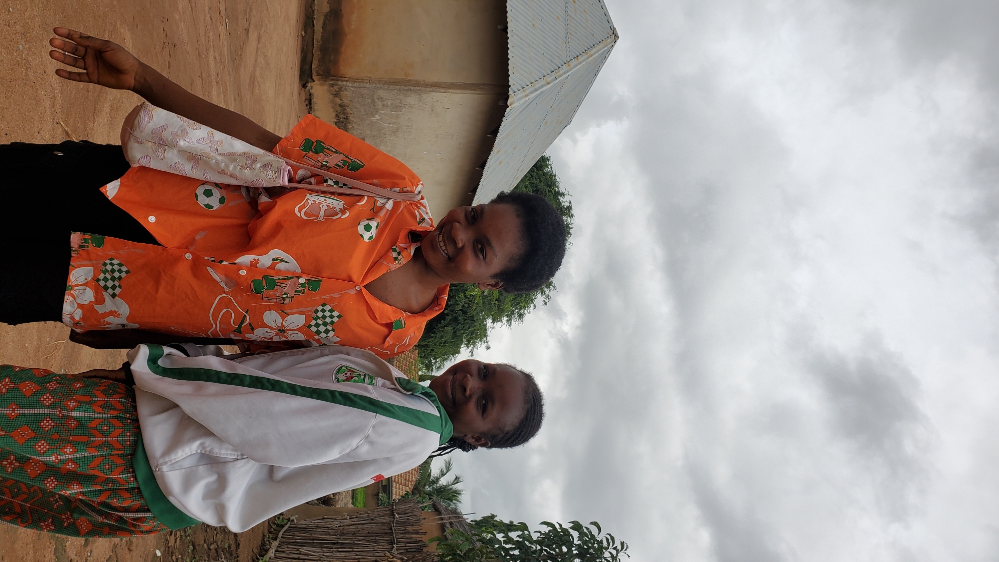 Two young women are standing next to each other outdoors, with a building and a fence in the background, likely in Jalingo, Nigeria. The woman on the left is wearing a bright orange short-sleeved button-up shirt with a pattern of soccer balls, flowers, and checkered squares. She has dark curly hair and is carrying a light-colored patterned bag over her shoulder. The woman on the right is wearing a white zip-up jacket with green trim and a green collar. Her hair is braided. They are both smiling and appear to be posing for a photo. The building behind them has a corrugated metal roof and beige walls. The ground is dirt, and there is vegetation and a rustic wooden fence visible. The sky is overcast.