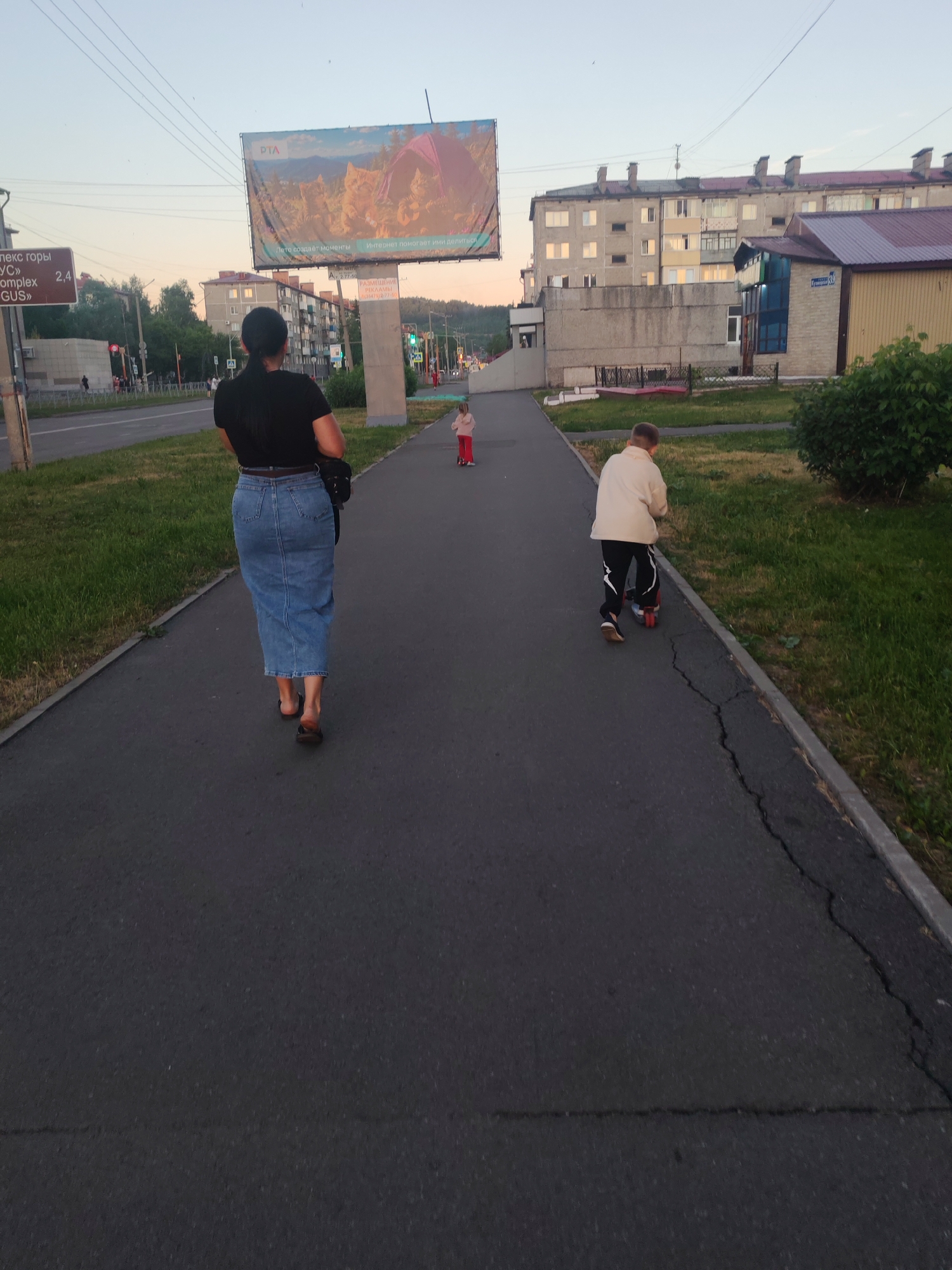 A full shot captures a paved walkway in Mezhdurechensk, Russia, with two children and an adult walking away from the camera. The walkway is bordered by a strip of grass on the left and a concrete curb on the right, with more grass and a dense green bush to the right. In the background, buildings with multiple stories and balconies are visible, along with a large billboard advertising a camping scene. The sky is a pale, hazy blue, suggesting either early morning or late afternoon.
The adult, a woman, is in the foreground, wearing a black t-shirt and a long denim skirt. She has dark hair pulled back in a ponytail and is carrying a black bag over her shoulder. She is wearing dark sandals and her feet are slightly spread apart, indicating she is walking.
A young boy on a scooter is positioned further down the walkway to the right. He wears a light-colored long-sleeved shirt and black pants with a white stripe down the side. He is leaning forward, appearing to be actively riding the scooter.
A younger child, likely a girl, is walking further ahead of the boy on the walkway. She is wearing a red long-sleeved shirt and red pants.
A directional sign with Russian text and numbers is visible to the left of the adult, indicating distances to specific locations. The overall scene depicts a tranquil urban environment with recreational activities.