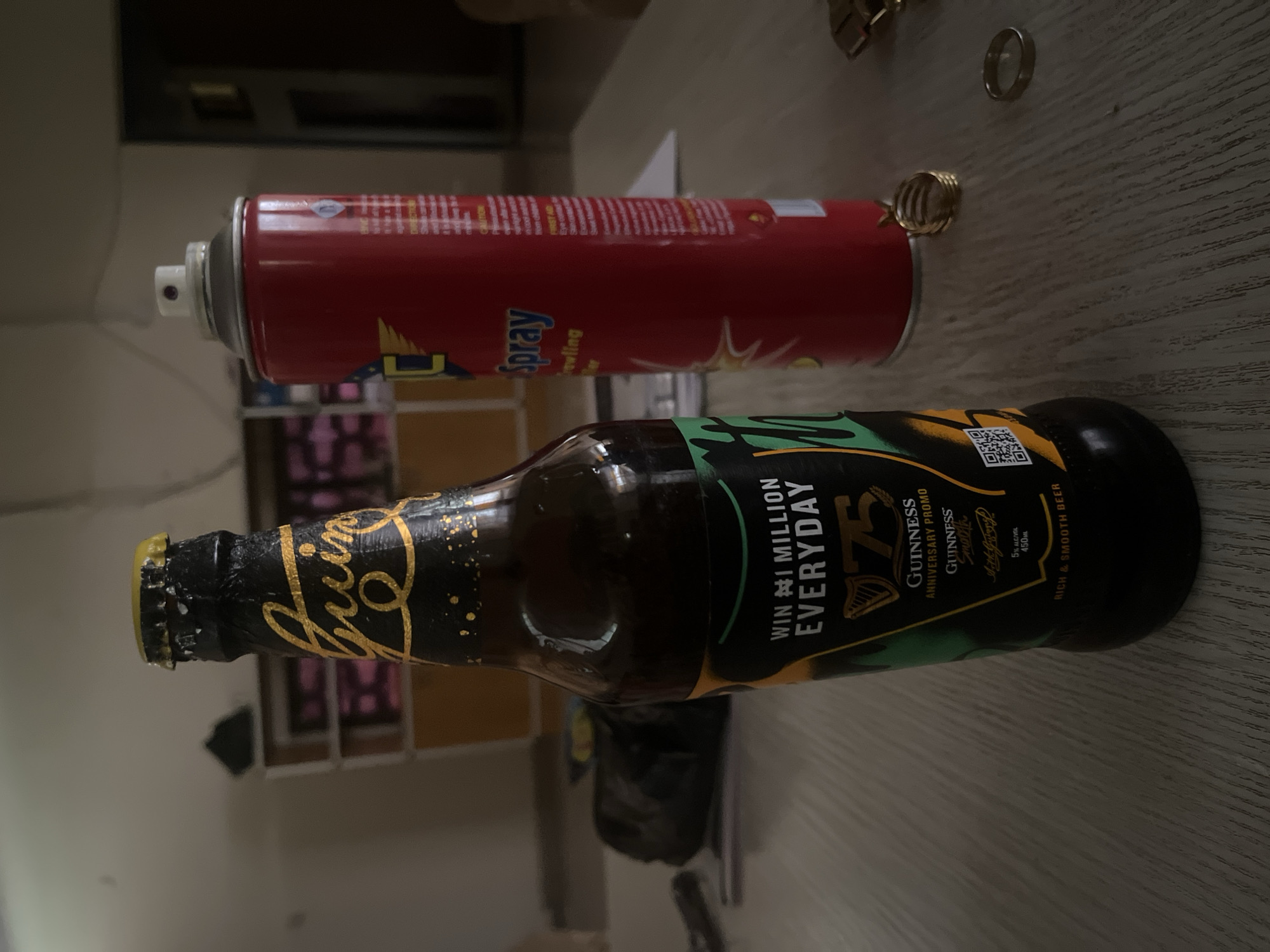 The image depicts a bottle of Guinness beer and a red spray can on a wooden table. The Guinness bottle has a black and gold label that reads "WIN #1 MILLION EVERYDAY" and "GUINNESS ANNIVERSARY PROMO". The spray can is red with a white nozzle and has "Spray" written on it in blue letters. Two rings, one gold and one silver, are also visible on the table. In the background, blurred shelves and a dark object are present. The scene appears to be indoors, possibly in a residential setting. There is no indication of the specific location within Zawan, Nigeria.