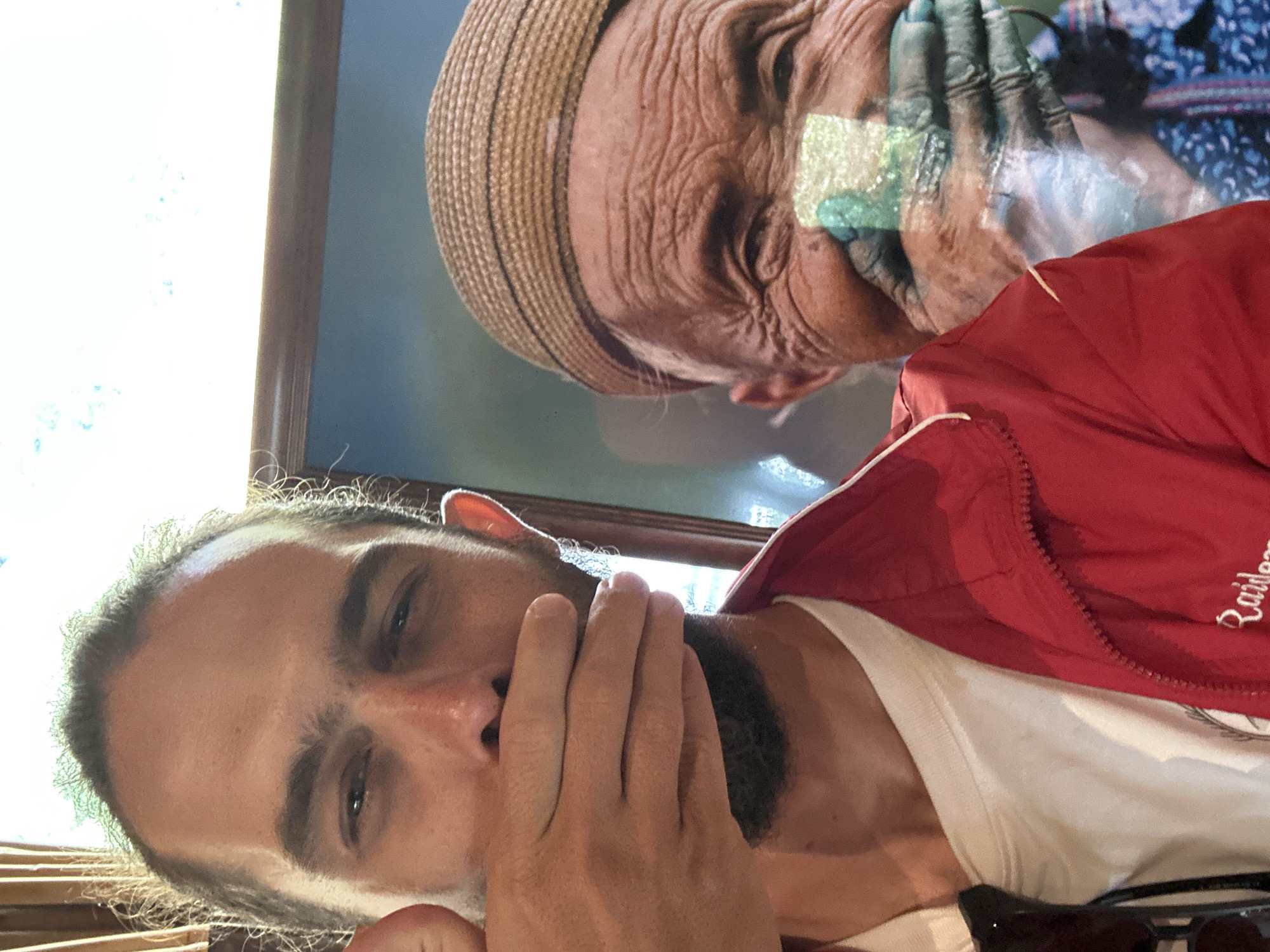 An individual is depicted in a selfie, posing in front of a framed portrait in Púng Luông, Vietnam. The foreground shows a light-skinned male with a beard and dark hair, wearing a white t-shirt and a red track jacket with "Raiders" partially visible on the chest. His left hand is raised, covering his mouth and lower nose, mirroring the subject in the background. His eyes are open, directed towards the camera. Behind him, a framed picture displays an elderly, heavily wrinkled individual wearing a light brown woven hat with dark stripes. This person also has their hands covering their mouth and displays a smile. A distinctive rectangular patch of light green and white pigment is visible on the subject's lower right cheek in the portrait. The background of the portrait is a soft blue, and the frame is dark brown. The lighting suggests an indoor setting with bright ambient light from behind the subject on the left.