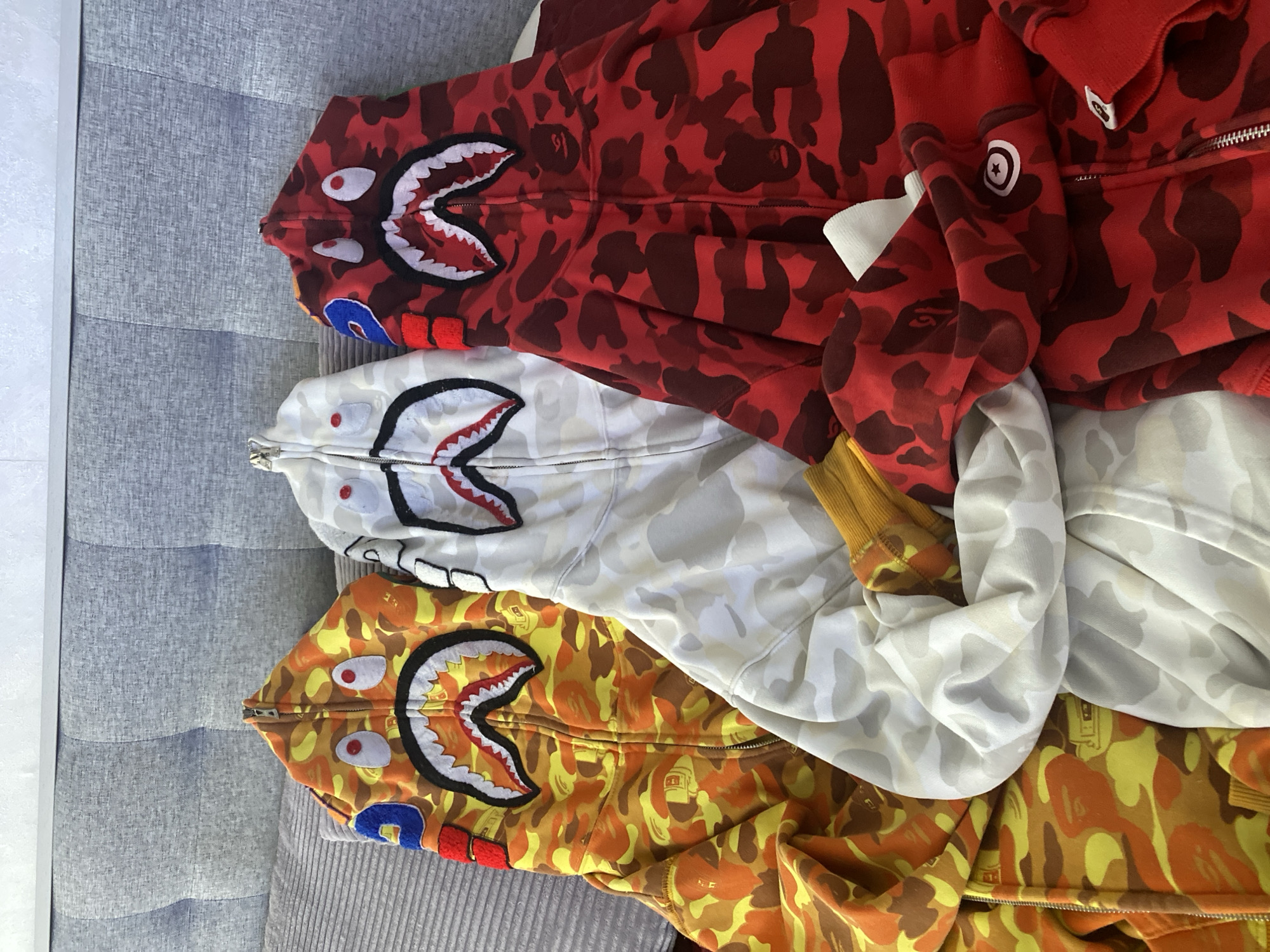 The image shows three BAPE shark hoodies stacked on a gray upholstered surface. The top hoodie is red camouflage, the middle is white camouflage, and the bottom is yellow camouflage. Each hoodie displays the shark face design on the hood. The location is not discernible from the image.