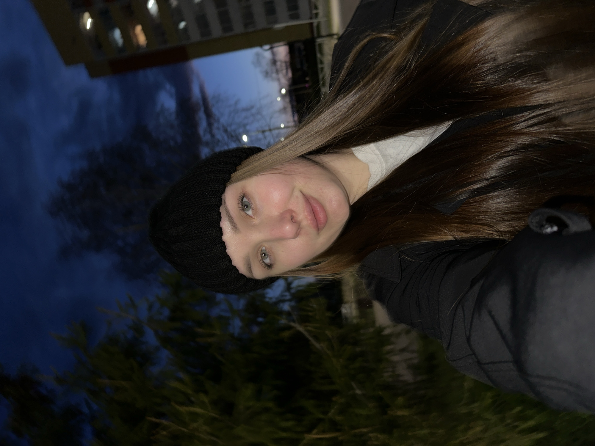 The image is a vertical-format selfie taken outdoors during the evening or night in Chaikovsky, Russia. The primary subject is a young woman, likely in her late teens or early twenties, positioned in the foreground. She has long, straight, dark brown hair, fair skin, and light-colored eyes (appearing green/blue). She is wearing a black knitted beanie and a black jacket over a white top, indicating cool weather. Her gaze is directed towards the camera with a neutral expression, showing a slight hint of a smile or smirk.
The background reveals a dark blue, twilight or nighttime sky. In the upper portion, a multi-story building, possibly a residential apartment block, is visible with several illuminated windows, confirming the time of day. In the lower background, dark green, dense evergreen trees or bushes are partially visible. The lighting is dim and ambient, with the woman's face appearing relatively well-lit, suggesting the use of a phone screen or flash. The image has a cool and subdued atmospheric quality. There is no visible text.
