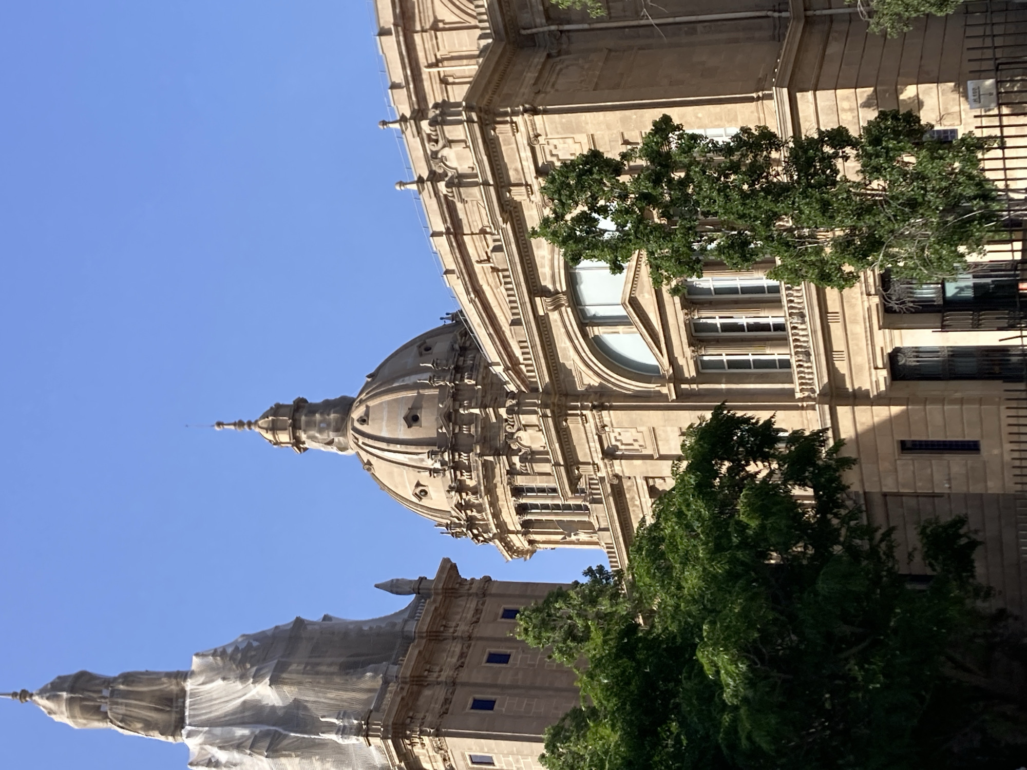 The image shows a detailed view of the Palau Nacional in Barcelona, Spain, against a clear blue sky. The building, made of stone, features a large central dome with intricate architectural details, including columns, arched windows, and decorative carvings. To the left, a tall tower is partially covered with scaffolding and protective sheeting, suggesting ongoing renovation work. Lush green trees in the foreground partially obscure the lower facade of the building, adding a natural element to the scene. The overall impression is one of grand architecture under a bright, sunny day. There are no people visible in the image.