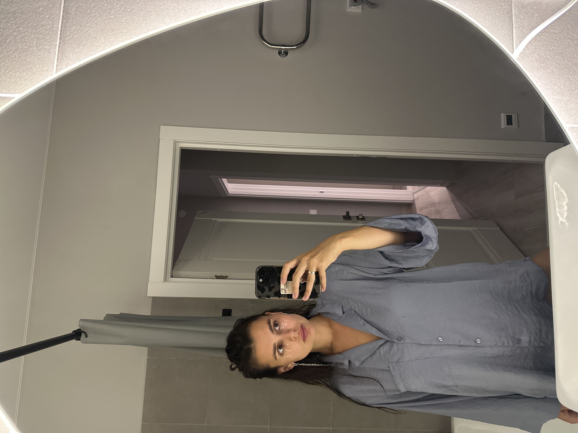 A young woman with long, dark hair tied up in a bun takes a mirror selfie in a bathroom. She is wearing a light blue, oversized pajama top and has glittery eye patches on. She is holding a black smartphone in her right hand, which is raised to take the picture. A silver earring hangs from her left ear. The bathroom has grey tiled walls, a shower curtain on a black rod, and a large, oval mirror with a light-up frame. A door is visible in the background, leading to a hallway. A white sink is in the foreground.