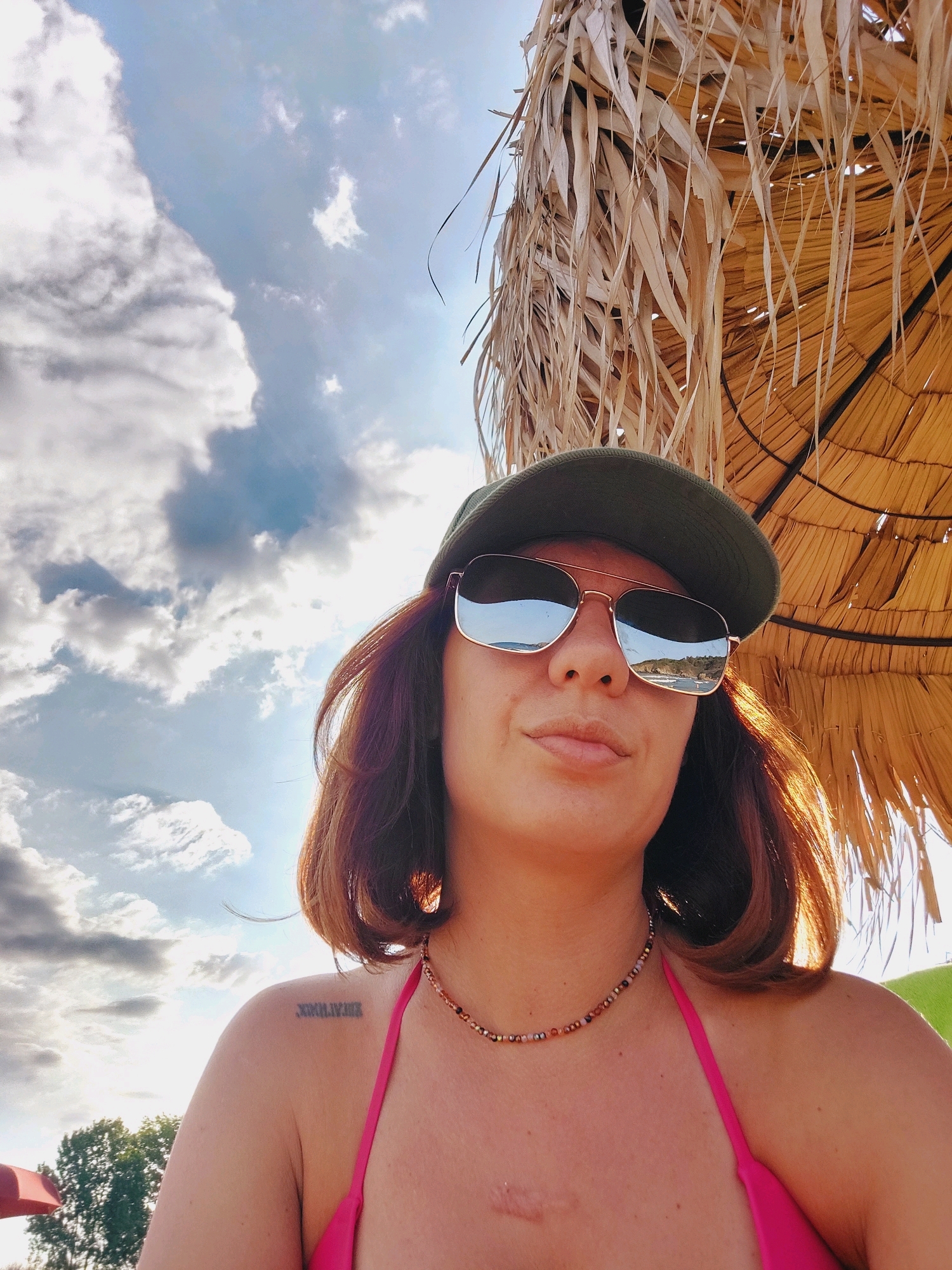 The image is a close-up, low-angle selfie of a woman with auburn hair and fair skin, taken outdoors. She is wearing a green baseball cap, aviator sunglasses, and a pink bikini top. A beaded necklace is visible around her neck. A faint scar is visible on her chest. She has a tattoo on her left shoulder, appearing to be Roman numerals. The background consists of a partly cloudy blue sky and a portion of a thatched umbrella. The sunglasses reflect a scene of a coastline with water and buildings. The overall setting suggests a beach or resort location, potentially in Sozopol, Bulgaria, given the context.