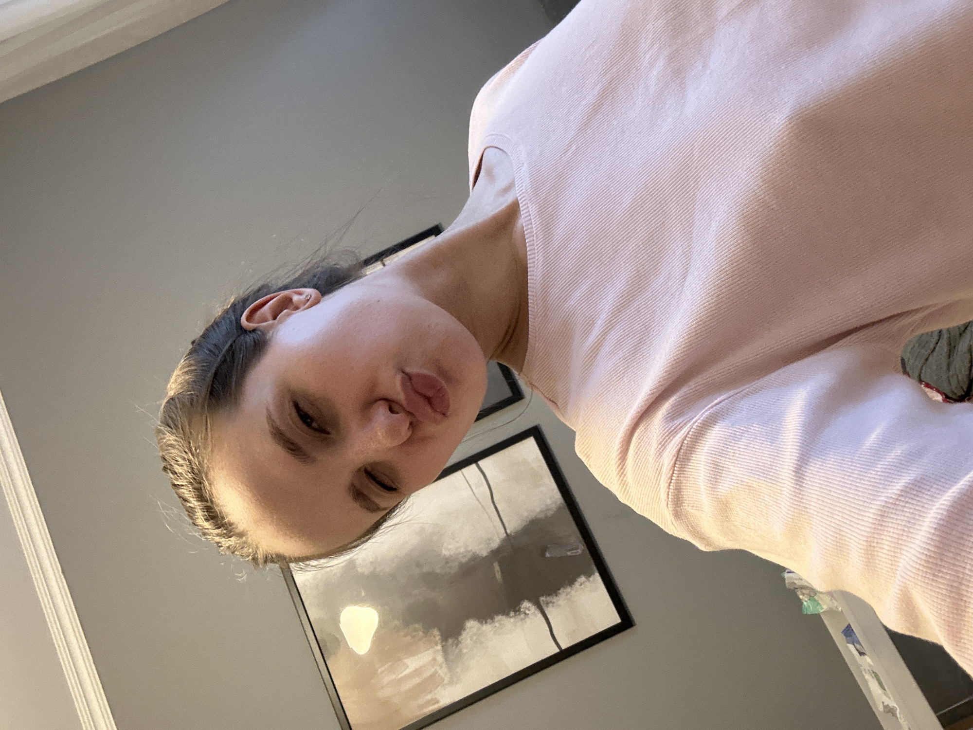 A person with fair skin and dark hair is wearing a pink long-sleeved shirt and looking down at the camera with pursed lips. The background includes a gray wall, a framed abstract painting, and a white-painted decorative molding on the left side. The photograph appears to have been taken indoors. The city of Khimki, Russia, cannot be determined from the provided image.