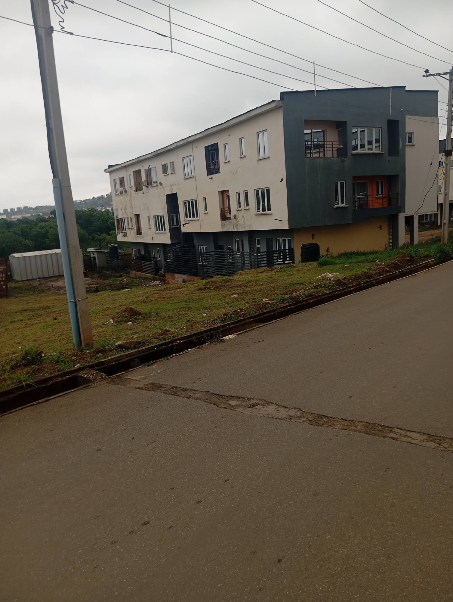 A street-level, eye-level, full shot captures a multi-story apartment building on an overcast day in Dutsen Alhaji, Nigeria. The building, painted in light beige and dark gray with an orange accent on the lower level, features multiple windows and balconies. The foreground is dominated by a sloping asphalt road with a cracked asphalt line running across it. To the left of the road, a grassy embankment borders the apartment building, which appears to be situated on a slightly elevated plot. A light gray utility pole with power lines extends upwards on the far left of the frame. The background shows a dense green landscape of trees and a hazy skyline of distant buildings. The scene is devoid of any people or animals.