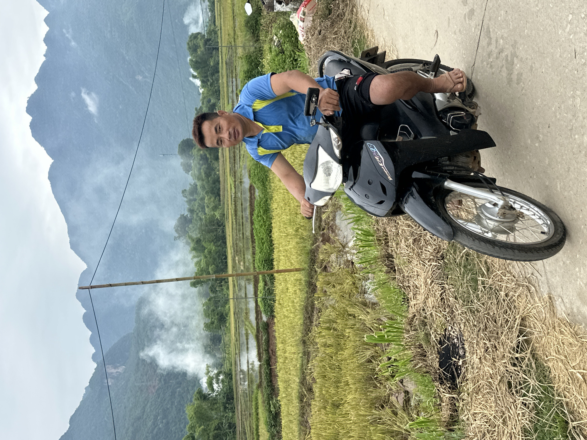 A middle-aged East Asian man, wearing a blue and yellow short-sleeved polo shirt and black shorts, rides a black motorcycle on a paved road. He is looking forward, with a neutral expression. The motorcycle has a silver front wheel and a black body. The man is wearing flip-flops.
The background shows a rural landscape in Mai Chau, Vietnam. There are rice paddies, some with harvested crops, and some with standing water. Green trees line the horizon. In the distance, mist partially obscures lush, green mountains. A tall, thin bamboo pole stands vertically to the left of the motorcycle. There is a visible power line stretching across the upper part of the image. The sky is overcast.
