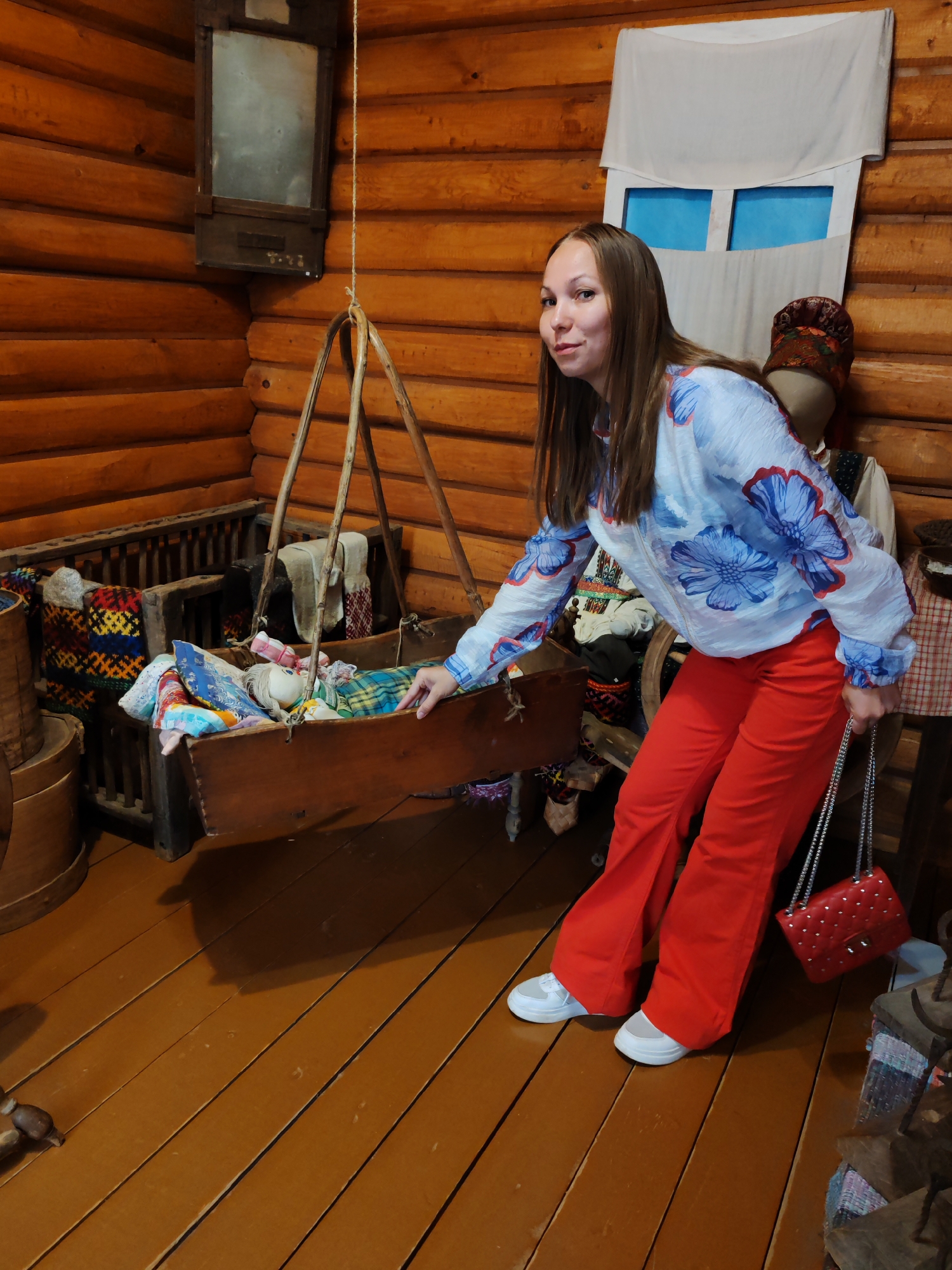 A woman stands in what appears to be a rustic, wooden-walled room, possibly a museum exhibit, in the village of Vizinga, Russia. She is wearing a light-colored, long-sleeved blouse with a blue floral print and bright red, flared pants. On her feet are white sneakers. She is holding a small, red quilted purse with a silver chain strap. Her left hand is extended and resting on the side of a wooden cradle suspended by ropes from the ceiling. The cradle is filled with colorful fabrics and what appears to be a doll. To her right, behind her, is a mannequin dressed in traditional clothing, including a patterned head covering. The room has wooden plank flooring and the walls are constructed from large, round logs. A small, framed mirror hangs on the wall above and to the left of the cradle. The overall atmosphere suggests a cultural or historical display.