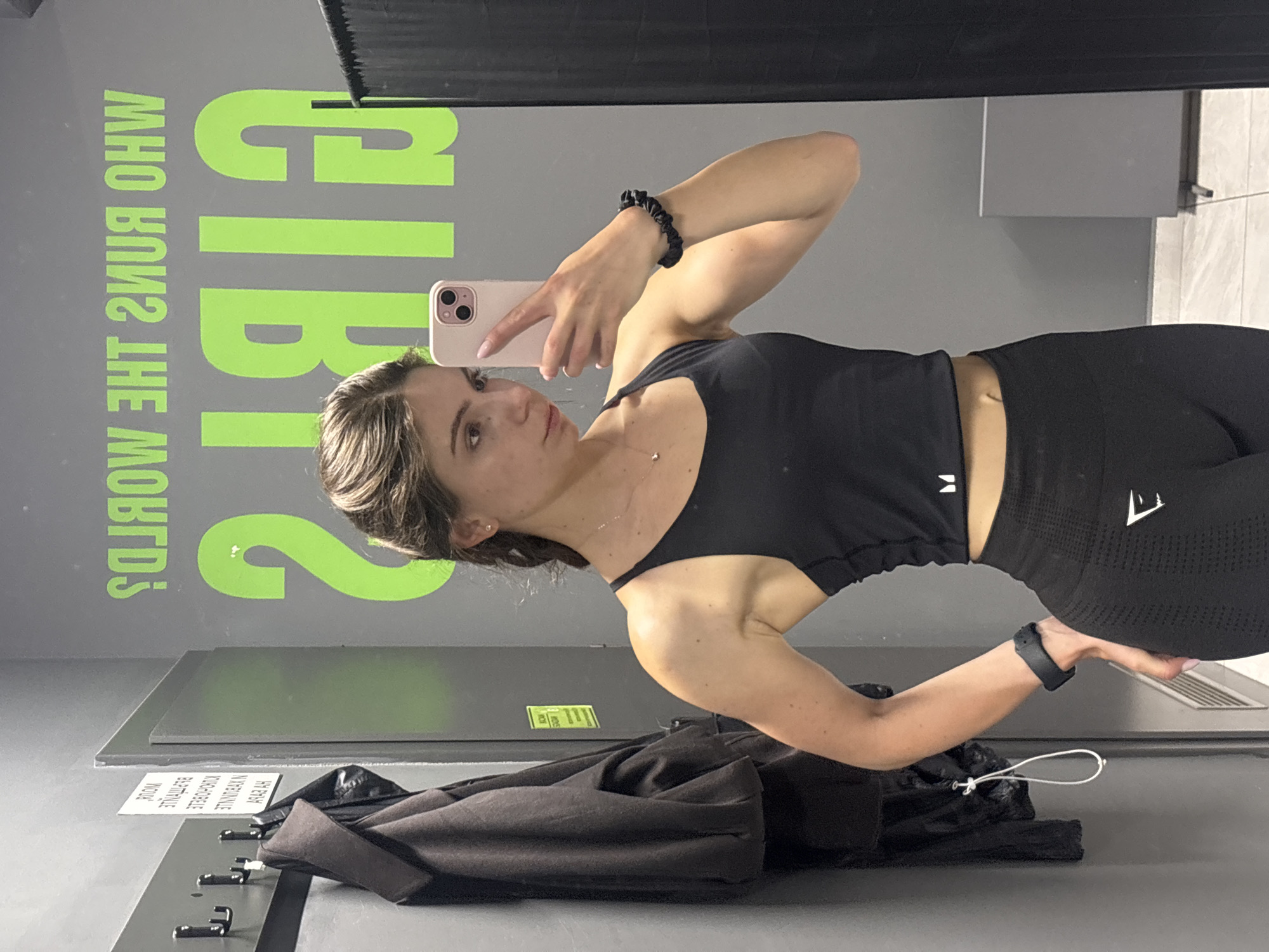 A woman is captured taking a mirror selfie in an indoor setting, likely a gym or fitness center. She stands with her body angled, holding a light pink smartphone to photograph her reflection. She has light brown hair, appears to be wearing minimal makeup, and is dressed in black athletic wear: a crop top and high-waisted leggings with a white logo on the left thigh. Accessories include a black scrunchie on her right wrist, a black fitness tracker on her left wrist, and a delicate necklace.
The background features a large mirror reflecting fluorescent green text that, when read correctly, states "GIRLS WHO RUN THE WORLD?". The floor is a dark gray or black mat. To the woman's right, a white laptop or tablet rests on the floor. To her left, a folded dark jacket or coat is placed on a shelf-like surface equipped with several black hooks. A piece of white paper with black text is also visible on the floor, featuring reversed words that include "ROOM" (from "VODD"), "BEZPRZELEWOWE", "KUCHENNE", and "NA ZAKUPY", suggesting Polish text. The scene is well-lit, characteristic of an indoor facility.