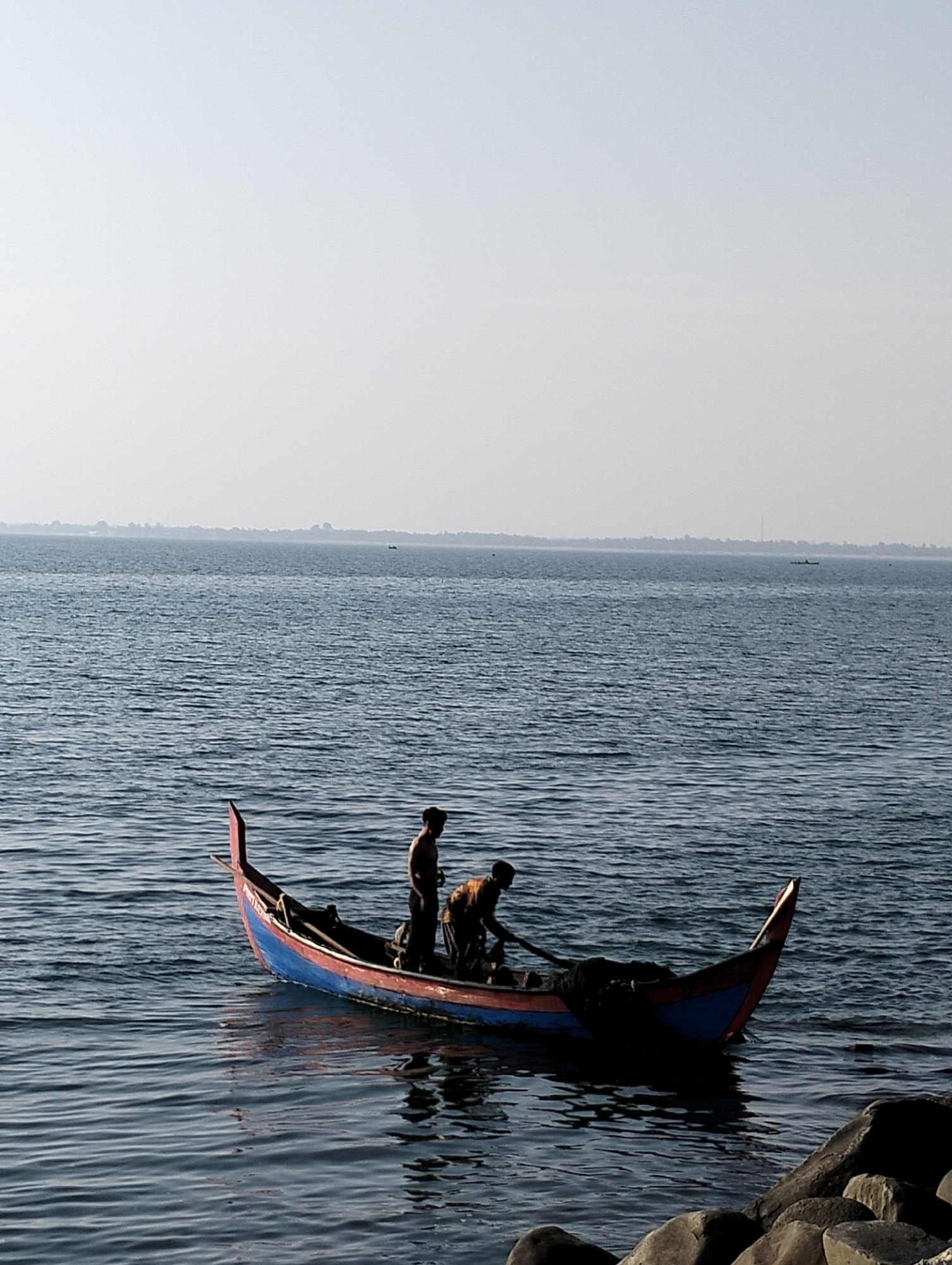 The image depicts a tranquil coastal scene in Lhokseumawe, Indonesia, featuring two men on a traditional wooden fishing boat. The boat, brightly painted with a blue hull and reddish-brown upper accents, rests on calm, rippling blue water. Its distinctive pointed bow and stern suggest a local design.
Two men are onboard, both appearing to be bare-chested or lightly clothed. One man stands near the stern, while the other is bent over towards the center of the boat, seemingly engaged with fishing nets or equipment visible within the vessel. Their activity suggests preparation for fishing or managing their catch.
The setting is an expansive body of water, likely a sea or bay, stretching to a distant, hazy horizon where a faint coastline or landmass is visible. The sky is bright and clear, indicating fair weather, possibly late morning or early afternoon, given the light conditions. In the foreground, dark rocks are partially visible, suggesting the boat is near the shore or a rocky bank. A smaller, indistinguishable boat can be seen as a faint silhouette further out on the water. The overall impression is one of quiet industry and daily life on the water in a peaceful maritime environment.