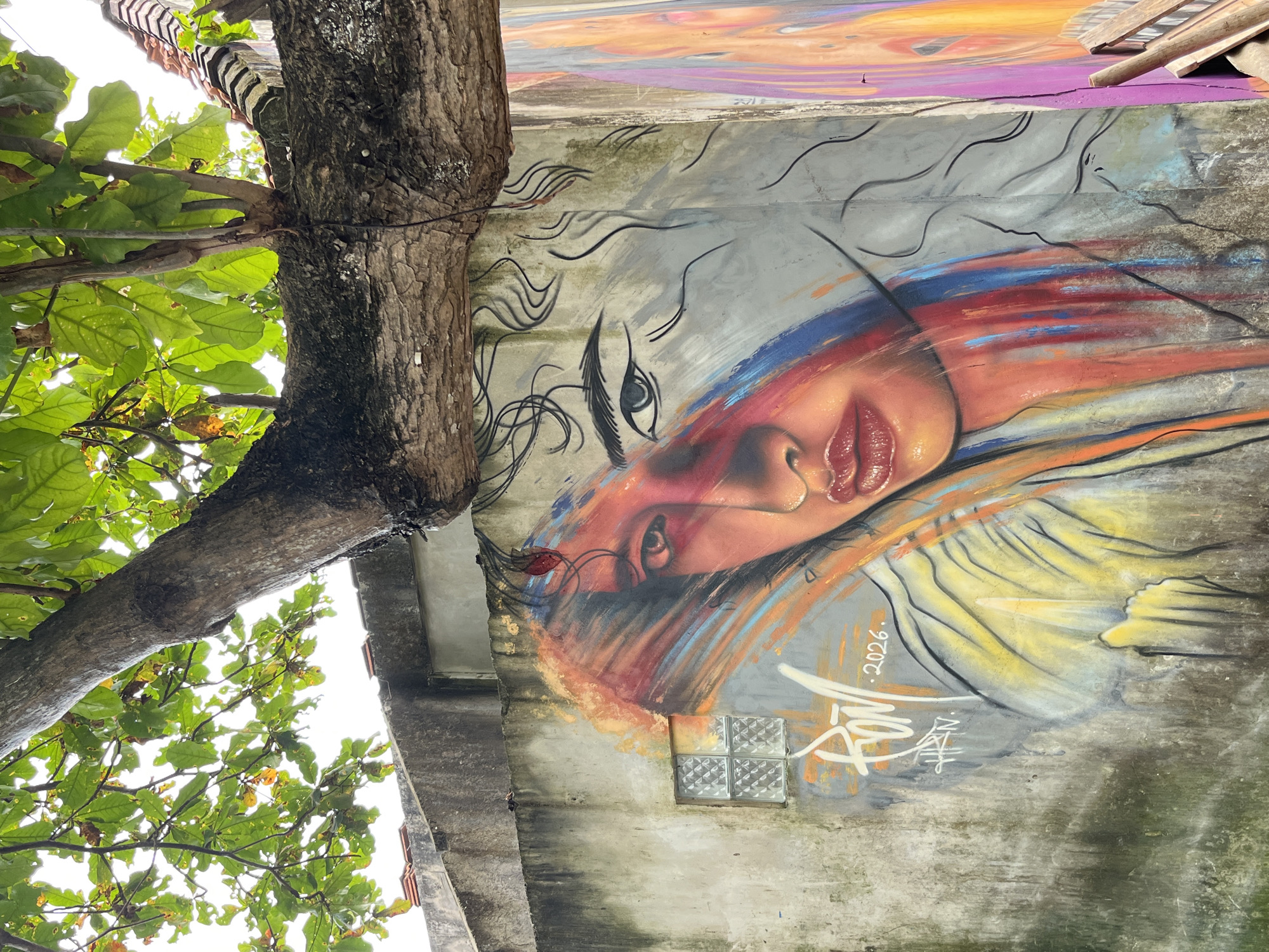 A large, colorful mural of a woman's face is painted on a weathered concrete wall in Kuta, Indonesia. The mural is partially obscured by a thick tree branch and lush green leaves overhead. The woman's face is depicted with bold strokes of red, orange, blue, and yellow paint, creating a dramatic effect. Her eye is a striking feature, with a detailed iris and dark eyelashes. Her lips are painted a glossy red, and her hair is rendered in swirling black lines. Below the mural, a signature and the year "2026" are visible. To the right of the mural, another partial mural of a face is visible, with vibrant pinks and oranges. A small, square window with frosted glass panes is set into the concrete wall to the left of the main mural. The overall scene suggests an urban setting with an artistic overlay, likely an outdoor public space.