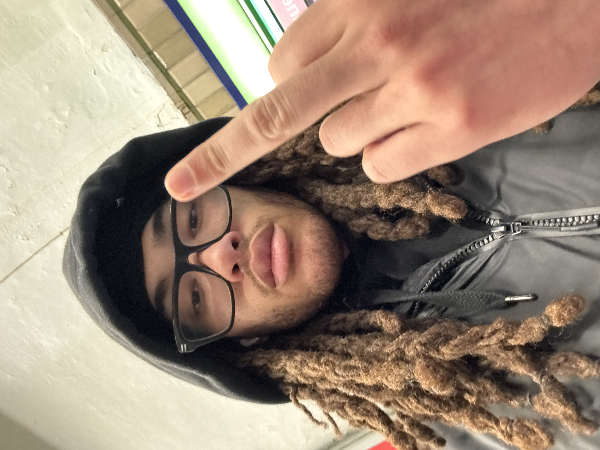 A medium shot of a man wearing a black hooded jacket with a closed silver zipper, black-framed glasses, and brown dreadlocks. He is holding up his index finger. He is located at the platform of a Paris Metro station.