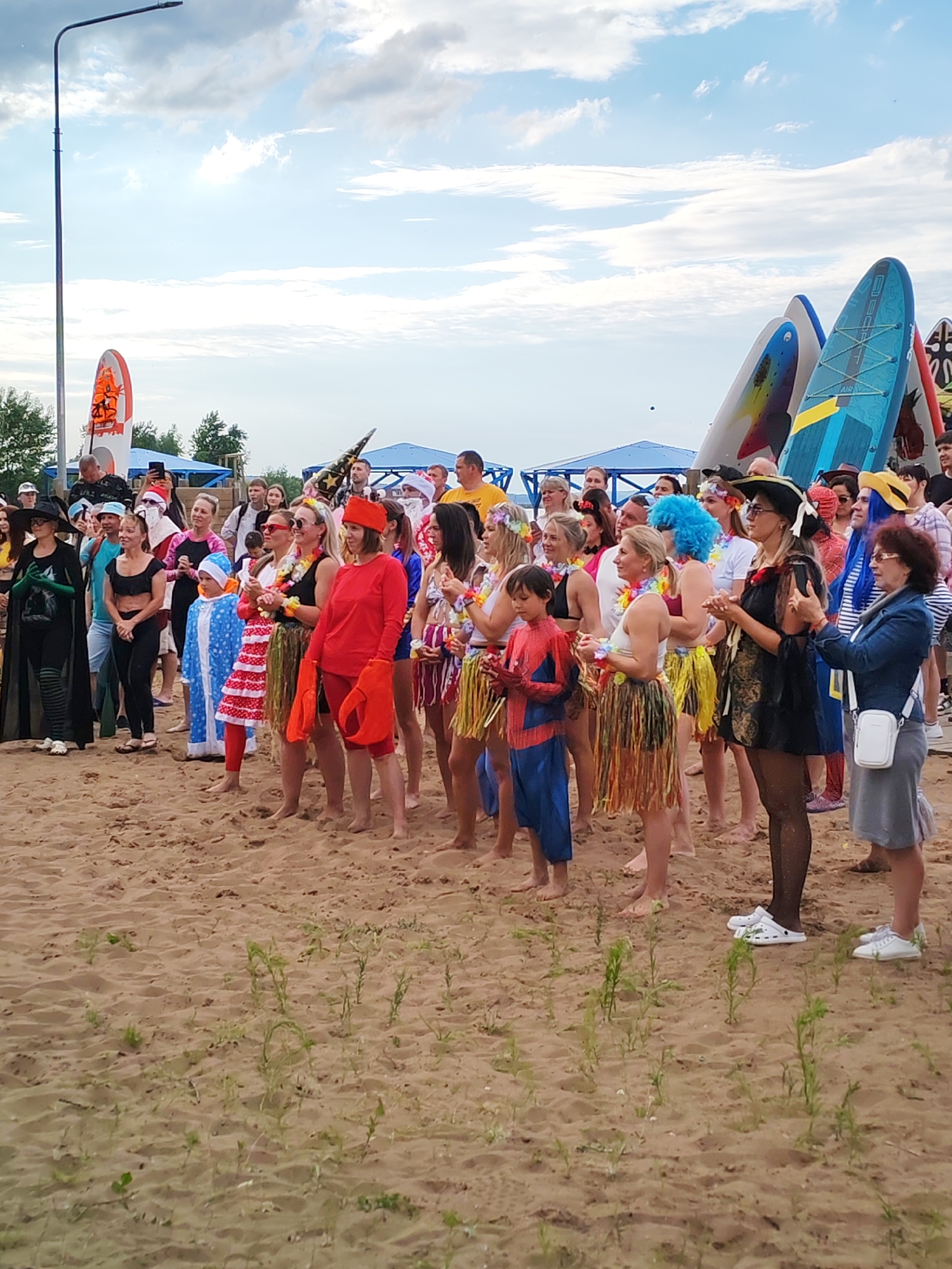 In Chaikovsky, Russia, a group of people are gathered on a sandy beach, some dressed in costumes. The group appears to be participating in an event or activity. Among the crowd, several individuals are wearing Hawaiian-themed attire, such as leis and grass skirts. One person is dressed as a crab, with large red claws. A child is dressed as Spider-Man. In the background, surfboards are visible, suggesting a connection to water sports. The sky is cloudy, and there is a lamppost on the left side of the frame. The overall atmosphere seems festive.