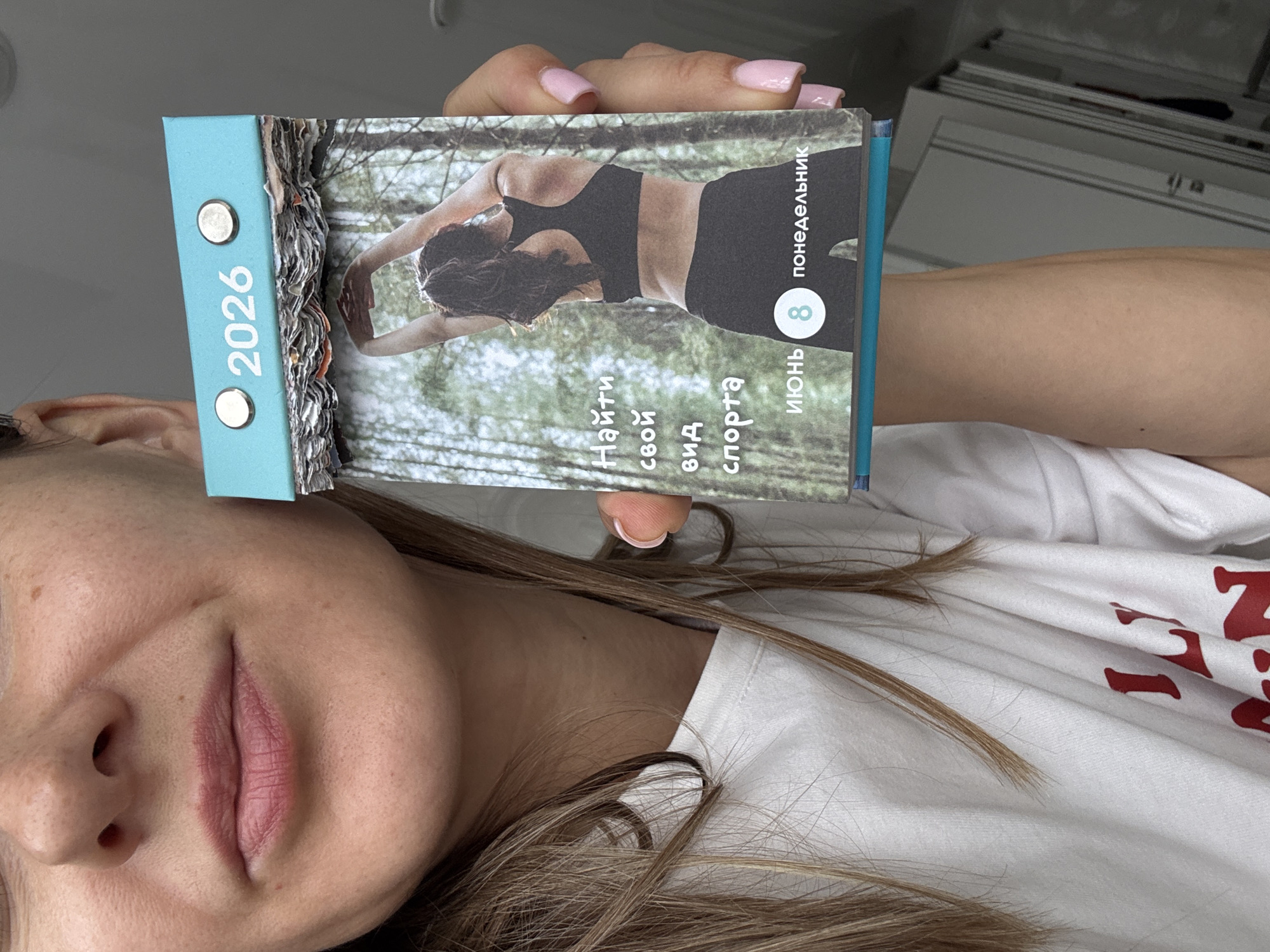 A woman is holding a desk calendar. The calendar has a turquoise cover with the year "2026" printed on it. Below the cover, the calendar shows a picture of a woman stretching outdoors, with trees in the background. The text on the calendar reads: "Найти свой вид спорта" which translates to "Find your sport." Below that, it says "ИЮНЬ 8 ПОНЕДЕЛЬНИК" which translates to "June 8 Monday." The woman's face, neck, and part of her chest are visible. She has blonde hair and is wearing a white t-shirt. Her fingernails are painted pink. The background appears to be an indoor setting with doors visible. No specific location in Sochi, Russia can be determined from the image.