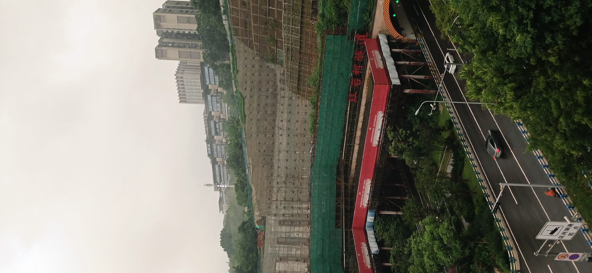 This detailed image captures an urban scene in Yuzhong District, Chongqing, China, under an overcast sky. In the background, several large, modern buildings with distinct architectural features, including one with a ribbed facade, crest a distant hill. A tall, slender tower, possibly for communication, is also visible on this elevated terrain. The midground is dominated by a substantial, engineered hillside secured by concrete retaining walls and sections covered with green mesh.
Below the hillside, a multi-tiered infrastructure system is prominent. An elevated structure, possibly an overpass, features large red banners with white Chinese characters on its facade. Beneath this, the entrance to the Hongyancun Tunnel ("红岩村隧道") is clearly visible, marked by a striped archway and overhead signage. Green traffic lights are active at the tunnel's opening.
In the foreground, a two-lane road, appearing wet, traverses the scene. Two vehicles are in motion: a dark sedan and a white car. A person on an orange scooter is also visible near the roadside. The road is bordered by blue and white barriers and flanked by abundant green trees and bushes. A street lamp stands prominently on the right side of the road, and a regulatory sign on the left indicates "区间测速 起点" (Section Speed Measurement Start Point).