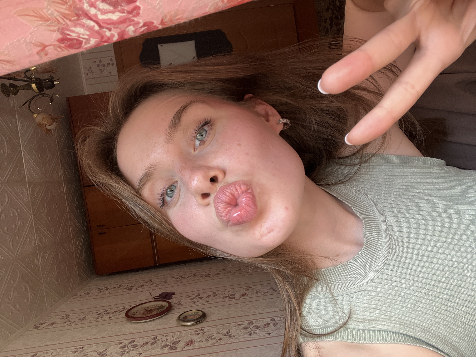 A young woman with light brown hair and blue eyes is taking a selfie in a room. She is puckering her lips and holding up two fingers in a peace sign with her right hand. She is wearing a light green ribbed sleeveless top. The background shows a wall with floral wallpaper and a wood-paneled ceiling. A clock is visible on the wall to her left. A chandelier hangs from the ceiling on the right side of the frame, partially obscured by a pink curtain with floral patterns. The overall impression is of a candid indoor photograph. The specific location in the city of Chaikovsky, Russia, cannot be determined from the visual information provided in the image.