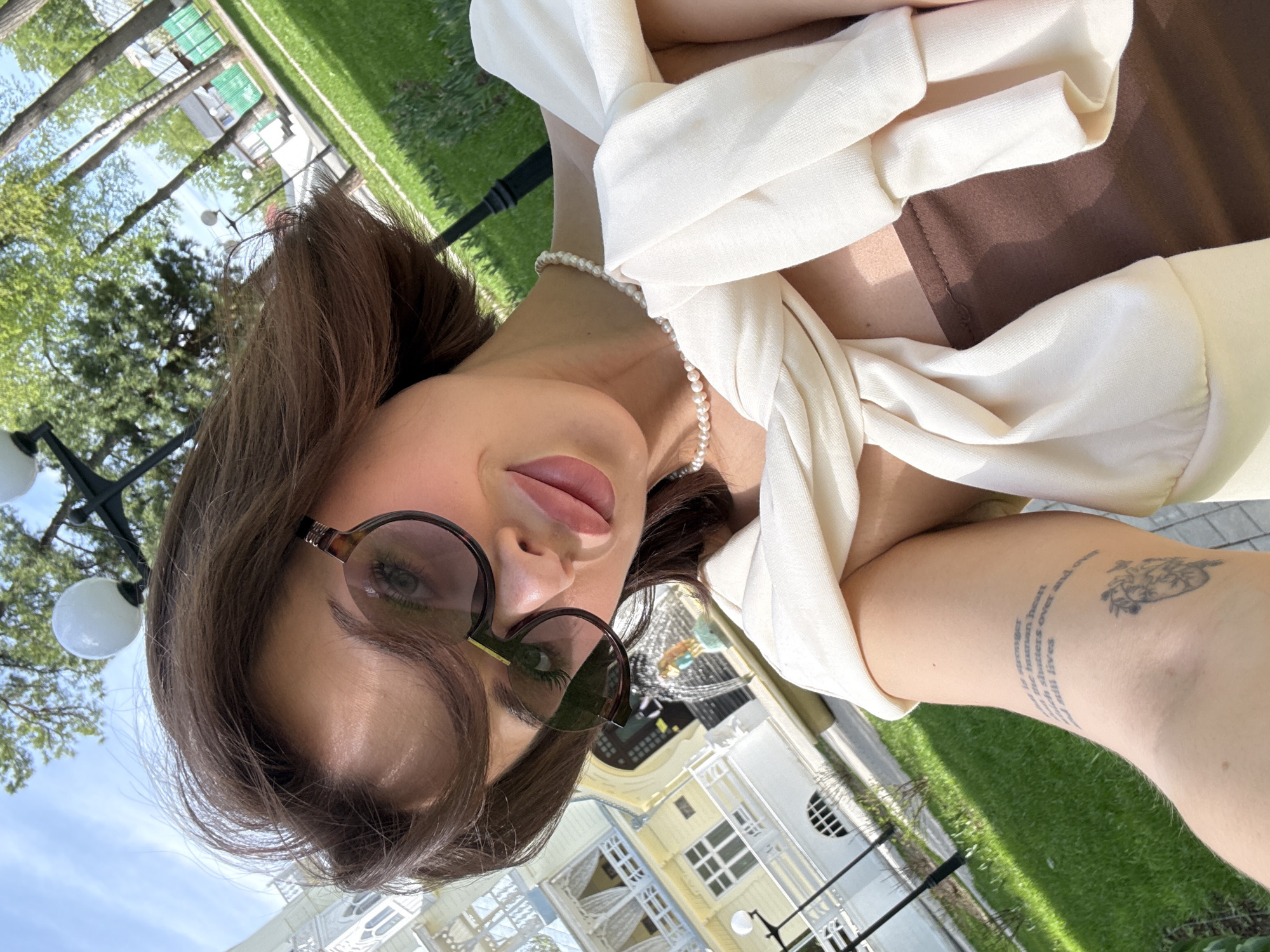 A woman with sunglasses and brown hair is taking a selfie outdoors. She is wearing a light-colored top with a brown underlayer and a pearl necklace. There is a tattoo on her arm. In the background, a building with white and yellow colors, along with trees and greenery, suggests an urban environment. The location is Sarapul, Russia.