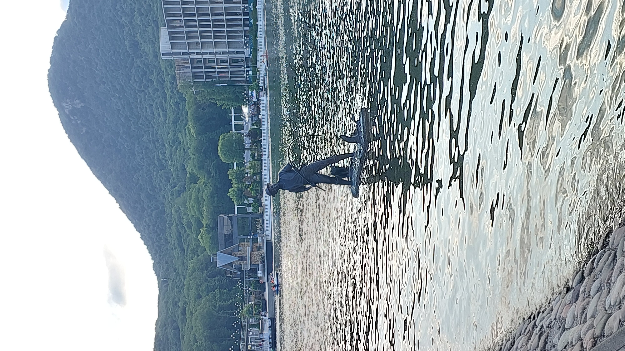 The image shows a statue of a man on a paddleboard in the water, with a rocky shoreline in the foreground. Behind the water, there is a tree-lined promenade with buildings and a large hill covered in trees. The sun is shining, creating reflections on the water. The statue appears to be made of dark metal and depicts a man in a fishing pose, holding a hoop. The overall scene suggests a peaceful day at a lake or reservoir, likely during the daytime given the bright sunlight and reflections. The location cues of a large hill covered in trees and a promenade suggest an outdoor recreational area. The image is oriented vertically.
