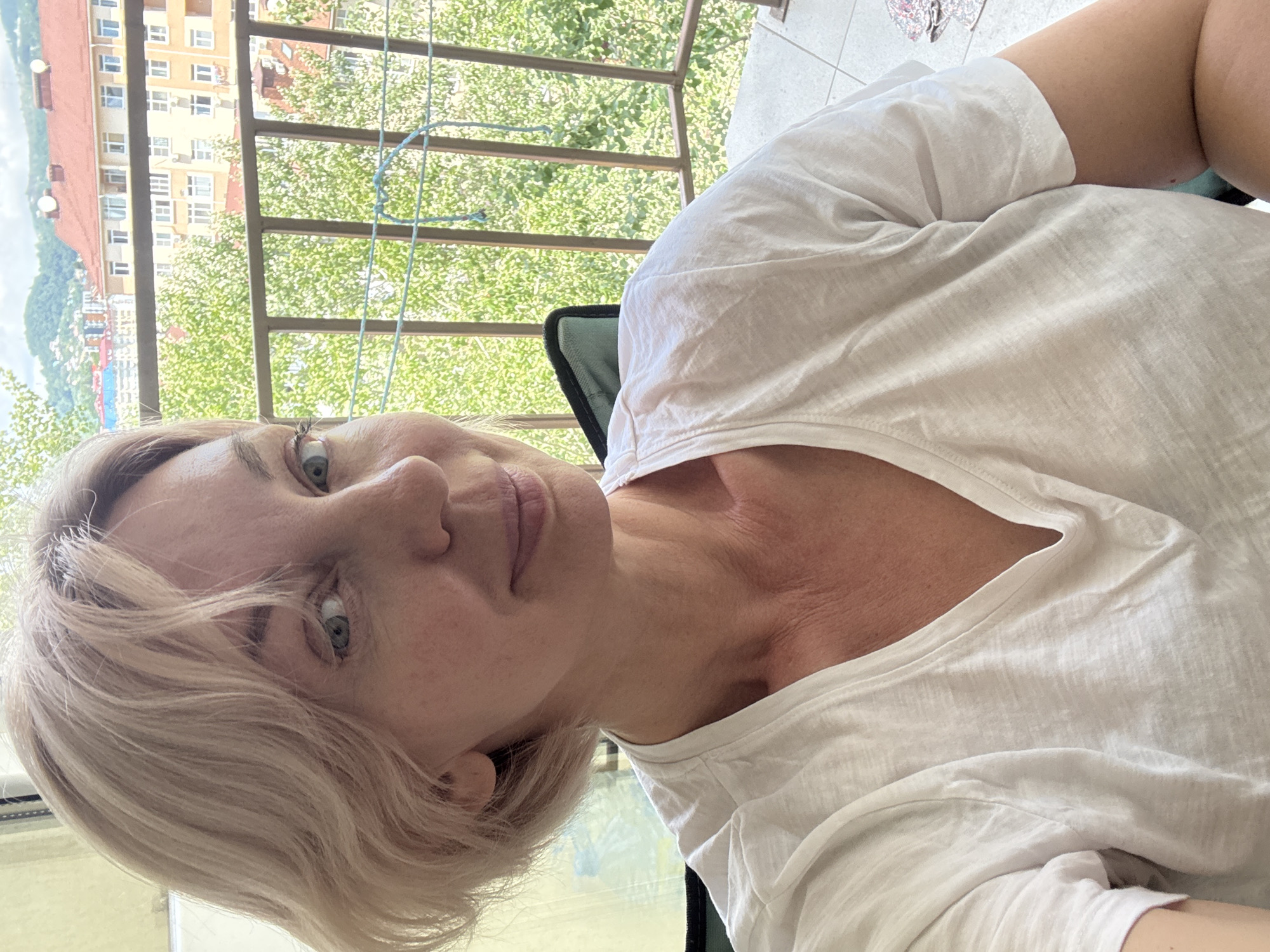A woman with short blonde hair is taking a selfie on a balcony. She is wearing a white v-neck t-shirt. In the background, there are trees, buildings, and mountains. The scene is likely located in Sochi, Russia, given the appearance of the architecture and the lush vegetation, which is consistent with the city's subtropical climate.