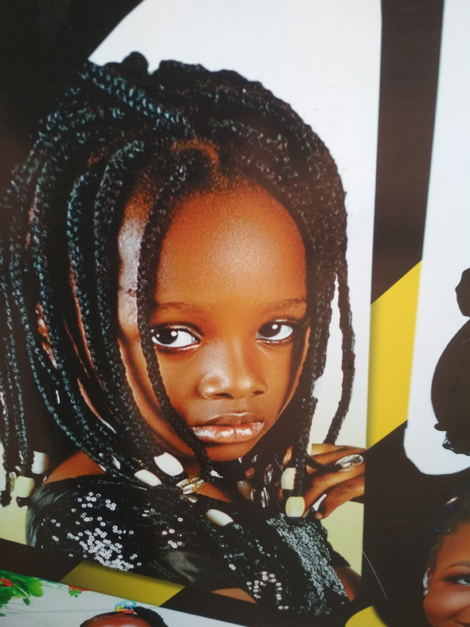 A close-up photograph shows a young, dark-skinned girl with braided hair. The braids are adorned with light-colored beads near the ends. She is wearing a black top with white sequins. The background appears to be part of a larger poster or image with black, white, and yellow sections, and partial views of other people. There is no indication of Jos, Nigeria.