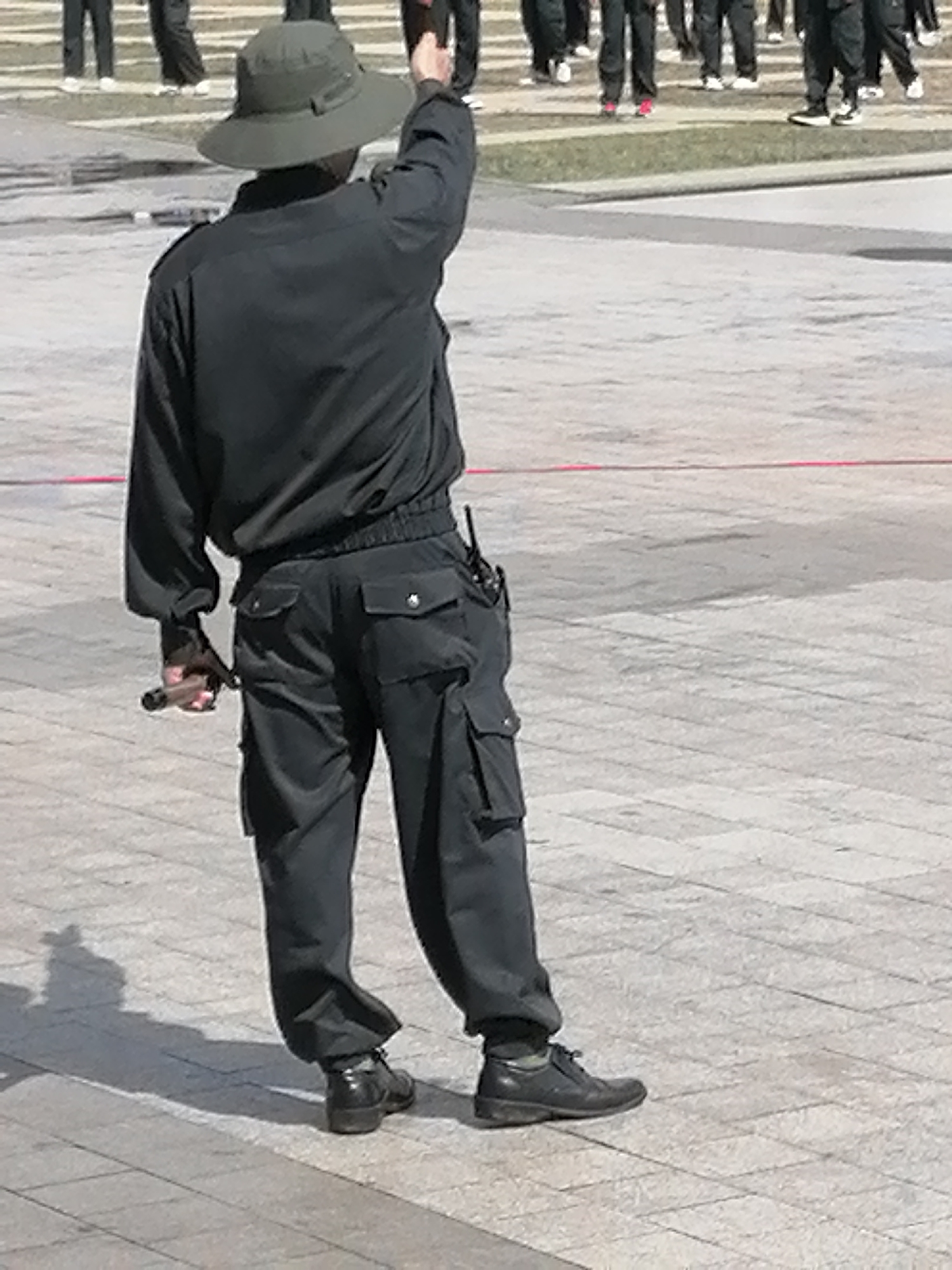 The image shows a person wearing a dark green wide-brimmed hat, a dark grey jacket with a collar, and dark grey cargo pants. The person is facing away from the camera, with their right arm raised and slightly bent, as if signaling or directing. They are holding a dark object in their right hand. The person is wearing dark, polished shoes. They are standing on a paved surface made of light grey square tiles, with faint red lines visible on the ground in the background. In the background, there are other individuals in dark clothing, some with white shoes, standing in a line. The environment appears to be an outdoor public space, possibly a plaza or parade ground. The lighting suggests it is daytime with direct sunlight, casting shadows on the ground. The image does not contain any specific landmarks or elements that would allow for the definitive identification of the location as Da Lat, Vietnam.