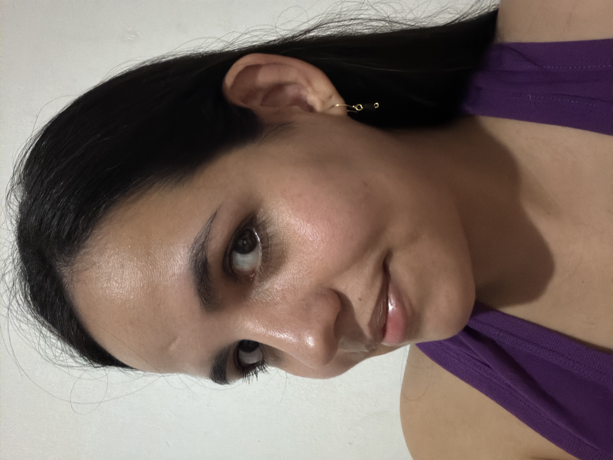 The image displays a close-up portrait of a female individual, captured indoors in Ciudad Guayana, Venezuela. The subject possesses long, dark hair, dark brown eyes, and a medium skin complexion. She is positioned with her head slightly turned to her left, looking directly into the camera with a subtle, upward curve at the corners of her lips. A faint horizontal line is present on her forehead. The subject is wearing a purple sleeveless garment. A gold-colored dangle earring, featuring two small, dark, circular elements, is visible on her right earlobe. The background is a plain, light-colored wall, lacking additional distinguishing features.