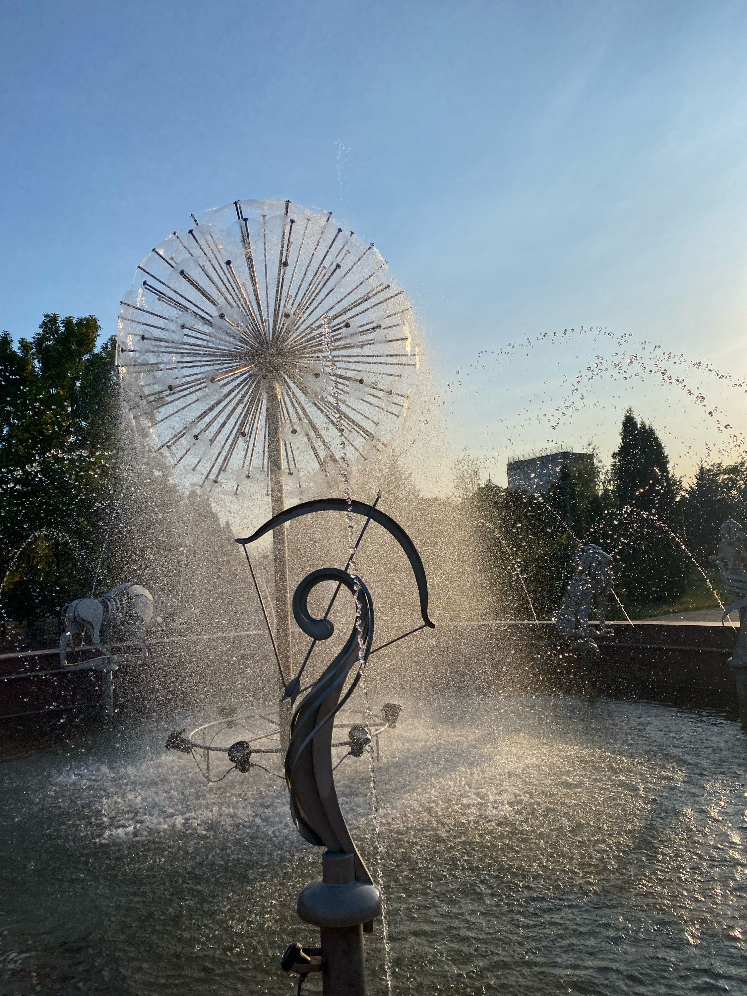 The image depicts a dandelion-shaped fountain with water spraying from its numerous metal rods. In the foreground, a metallic sculpture resembling a spiral and a bow-like structure is visible. The fountain is situated in a pool of water with a rough, textured surface. In the background, trees, a building, and statues of animals can be observed. The scene is bathed in the warm light of what appears to be late afternoon or early morning. This fountain is located in the "La Izvor" park in Chișinău, Moldova.