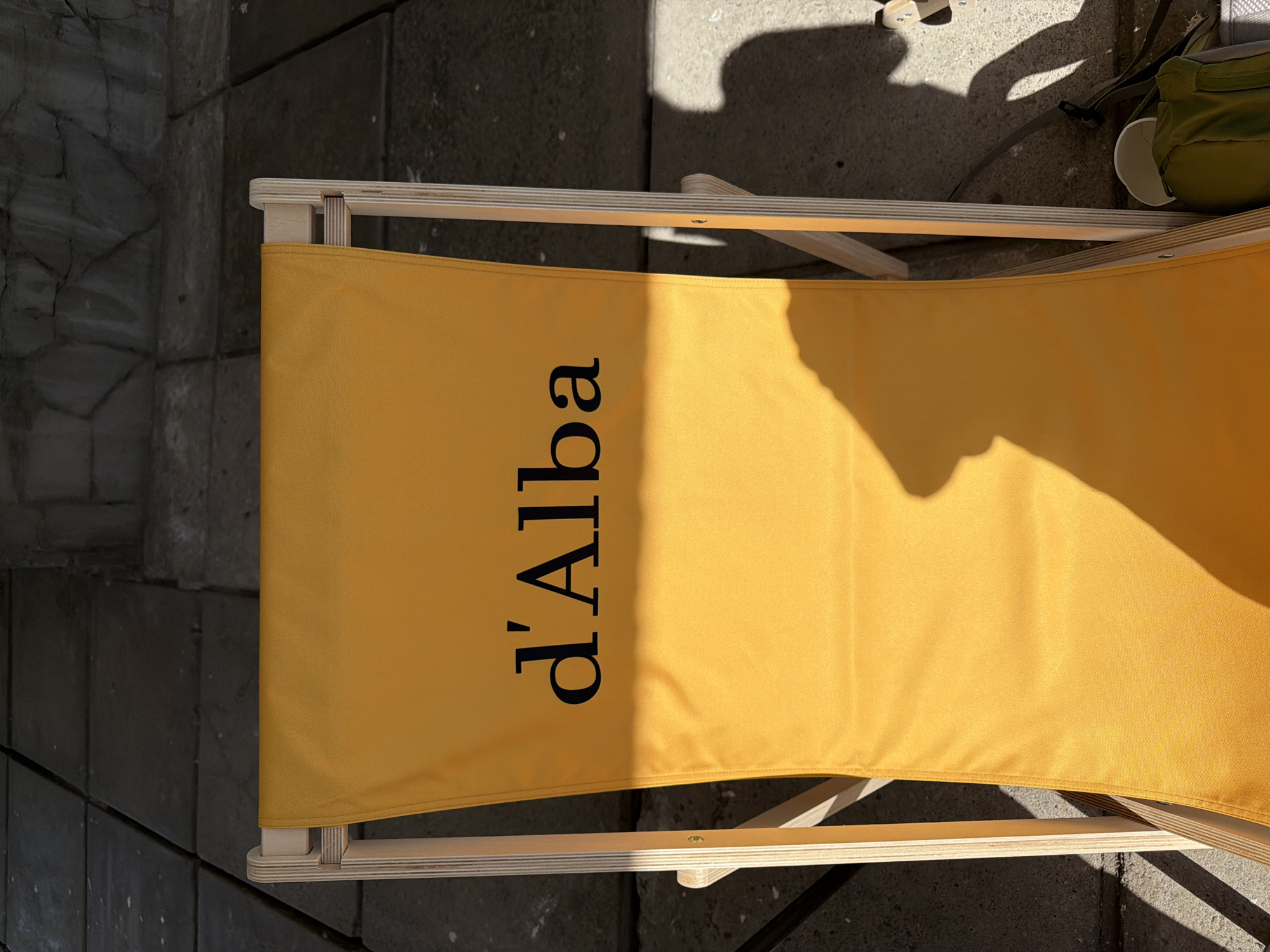 A high-angle outdoor image depicts a yellow fabric deck chair with a light-colored plywood frame, situated on a dark gray paved surface. The word "d'Alba" is printed in black serif font on the yellow fabric backrest. Strong sunlight illuminates the scene from the left, casting a distinct shadow, consistent with a human head and upper body, across the right side of the chair and the adjacent paving stones. The paved ground consists of rectangular, irregularly textured stone or concrete slabs. Behind the chair, a dark, textured wall or raised stone surface is visible. To the right of the chair, partially obscured by shadow on the paved ground, a green bag-like object and a white cup are present. No individuals are directly visible within the frame, only their cast shadow. The scene is located in Saint Petersburg, Russia.