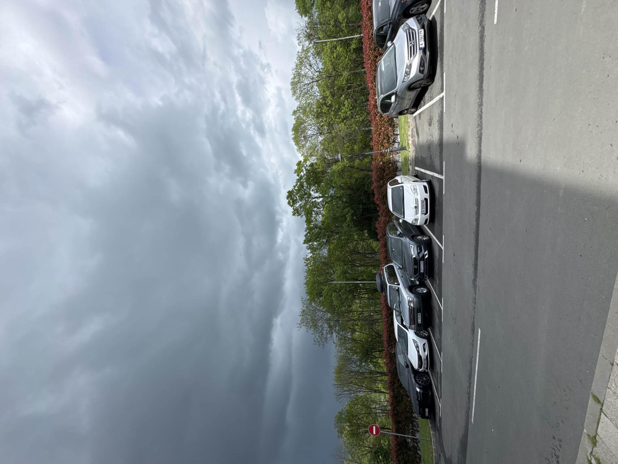 The image depicts a parking lot adjacent to a landscaped area with trees and shrubs. The sky is dominated by a large, dark storm cloud formation.
Several cars are parked in the lot, including a white car with license plate "A 5762 BX", a silver car with license plate "A 1067 PK", and a black car with license plate "JP4455C". The cars are neatly parked in designated parking spaces.
A "No Entry" sign is visible in the lower part of the image.
The location is likely in Burgas, Bulgaria due to the license plates and other details visible in the picture.