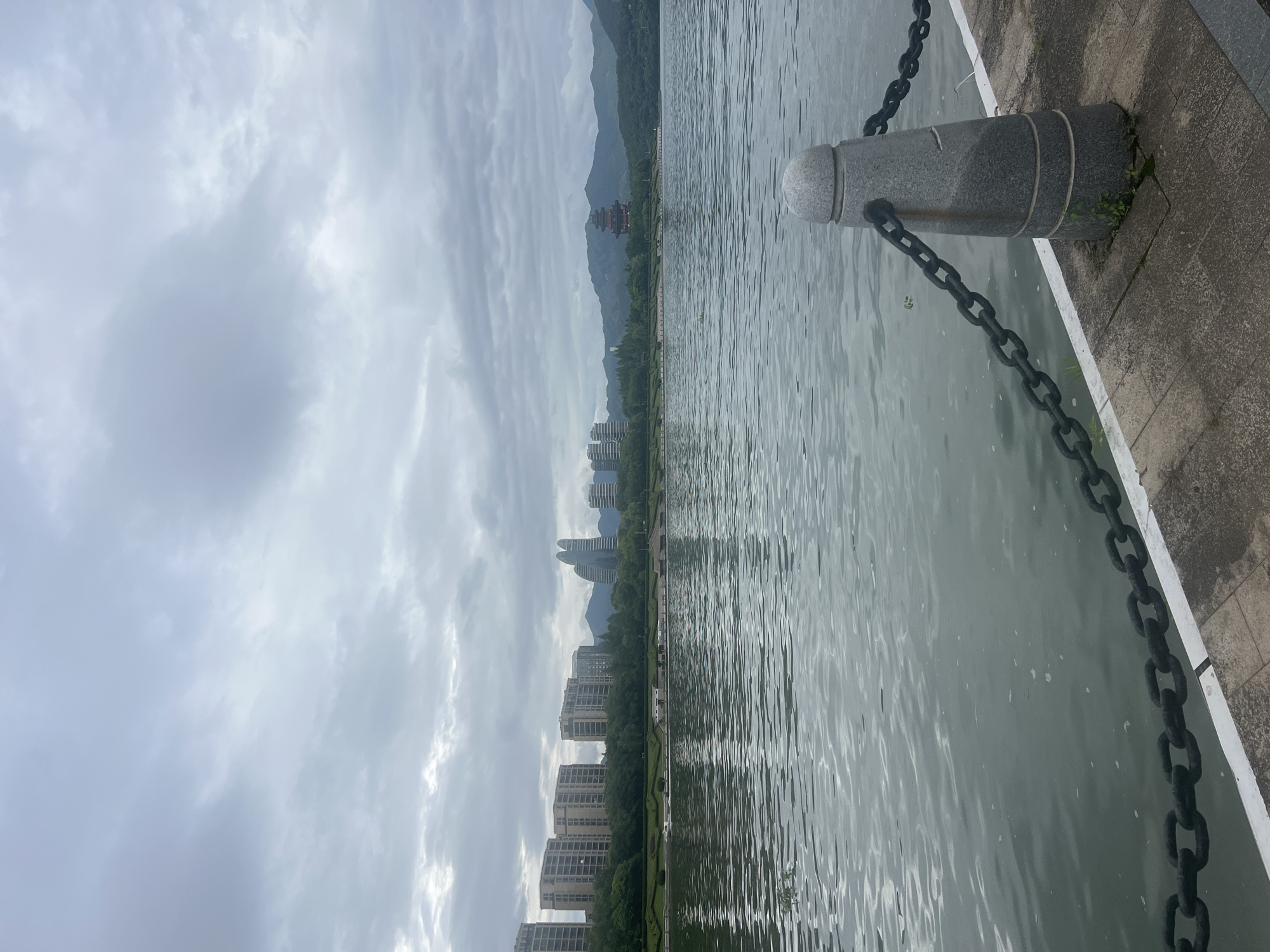 The image depicts a waterfront scene in Liandu District, China. In the foreground, a concrete pathway lines the edge of a body of water. A grey granite bollard with horizontal grooves and a rounded top is situated on the pathway, connected to a heavy black metal chain that extends along the water's edge. The water is calm, with ripples creating a textured surface that reflects the overcast sky.
Across the water, the opposite bank features a line of modern apartment buildings with multiple stories. Behind the buildings, a lush green treeline separates the urban development from rolling hills. Further in the distance, a multi-tiered pagoda-style building with a dark, ornate roof stands prominently against the backdrop of the hills. The sky is heavily clouded, contributing to a muted, diffused light. No people are visible in the image.