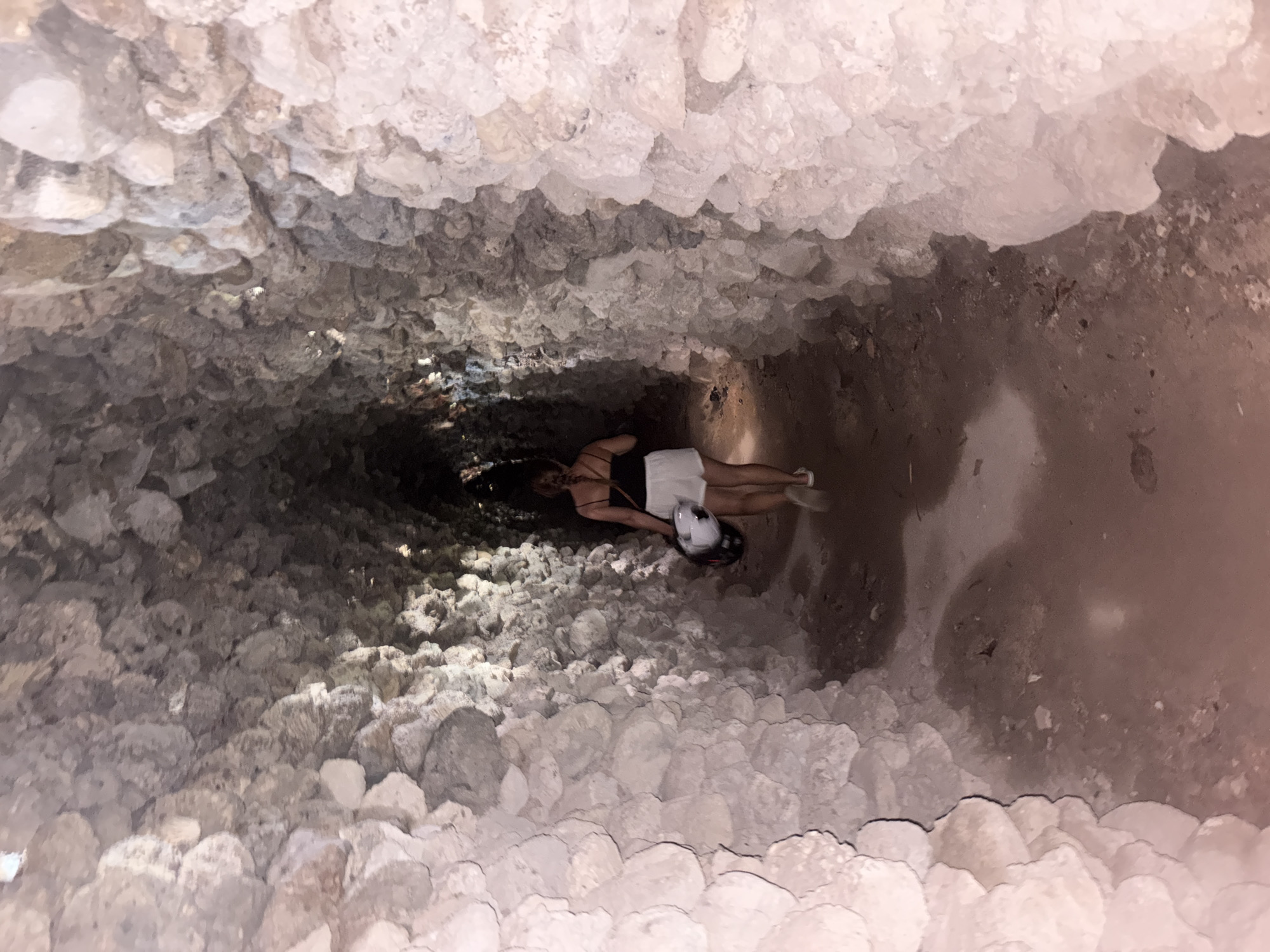 The image captures a top-down view of a person inside a deep, narrow shaft or cave. The walls of the shaft are extensively covered in light-colored, rough, and irregular formations, resembling crystallized deposits or small, knobby stalactites, creating a highly textured environment. The lighting is dim, suggesting an enclosed subterranean space or nighttime, with light appearing to come from the opening above or an artificial source near the subject.
A woman is visible lying on her back near the bottom of the shaft, looking upwards towards the camera. She has dark hair and is dressed in a black top or swimsuit and white shorts, with light-colored shoes. A white helmet with dark accents is positioned near her head or chest. Her posture suggests she is either resting, in the process of descending or ascending, or exploring the depths of the shaft.
The visible floor beneath the woman appears to be dark earth or rock. The overall scene conveys a sense of exploration within a unique geological feature or a man-made subterranean structure. Given the context of Cam Ranh, Vietnam, this could be a cave system, a deep well, or an old tunnel known for its distinct mineral formations. No text is visible in the image.