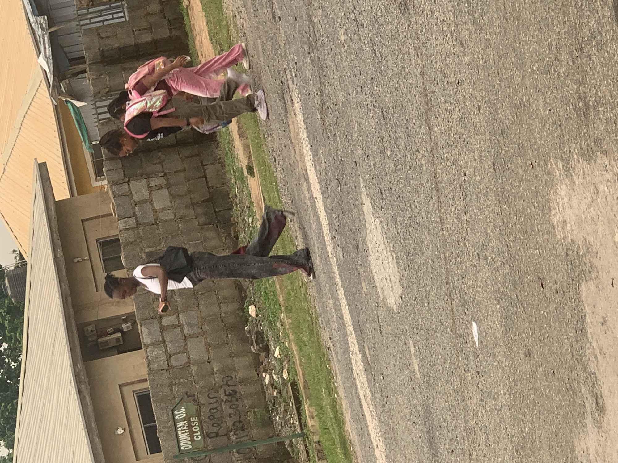 The image shows two people walking on a street in Abuja, Nigeria. The street is paved with asphalt and has a white line marking the edge. On the left side of the street, there is a building made of concrete blocks and a corrugated iron roof. There is also a patch of green grass and some debris.
The first person, from the top of the image, is wearing pink tracksuits and carrying a backpack. They are walking away from the camera. The second person is wearing a white t-shirt and dark pants, with a black backpack. They are walking towards the camera. Both individuals appear to be young.
The scene suggests a daytime setting, likely a warm day given the clothing. There are no obvious weather indicators, but the lighting implies it's not very early or late in the day. The building appears to be a residential or commercial structure, possibly a shop, given the sign visible on the wall.
On the wall of the building, there is some graffiti. One sign reads "ODUNTAN O.C. CLOSE" and another appears to have "Repair" written on it. Below "Repair", there is a number which is partially visible as "1X23035". The overall impression is that of a typical street scene in a Nigerian city.
