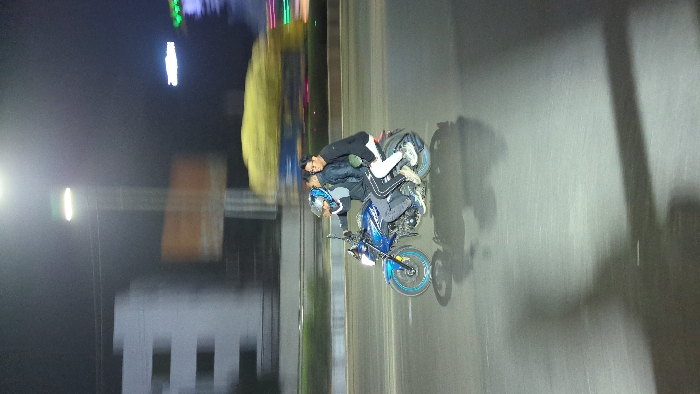 Two men are on a motorcycle performing a stunt on a street. The rider wears a helmet and dark clothing, while the passenger wears a jacket and light-colored pants and shoes. The motorcycle is blue with a custom wheel. The background shows a street at night with buildings and lights. The image is rotated and blurred.